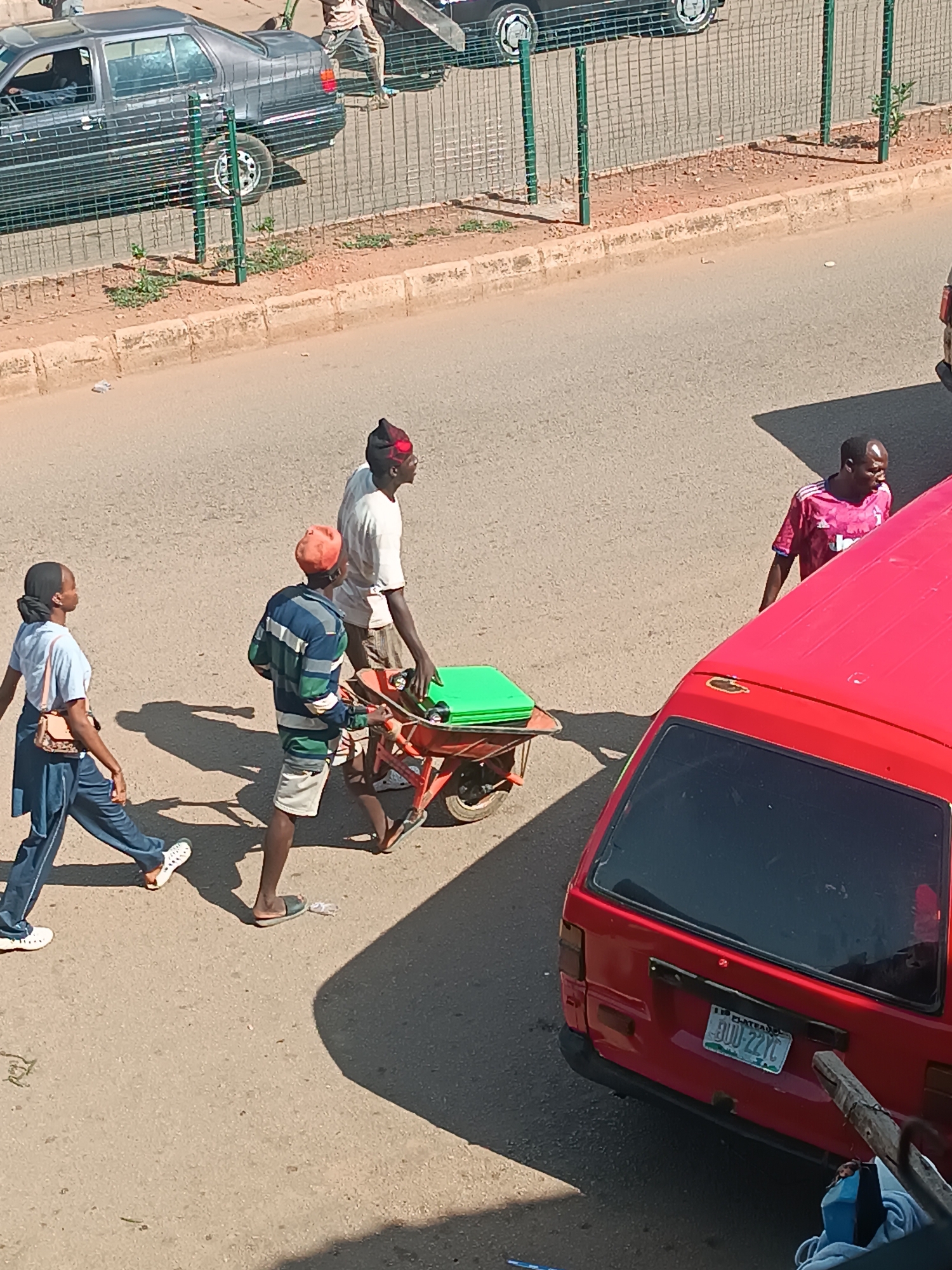 The image shows a street in Bukuru, Nigeria. A woman in a blue outfit walks on the left side of the frame. A man in a striped shirt and orange hat is assisting a man in a white shirt and black hat in pushing a wheelbarrow loaded with green objects. Another man in a red and pink jersey walks behind the wheelbarrow. A red van is parked on the right side of the frame. A gray car is parked on the other side of the street, behind a wire fence.