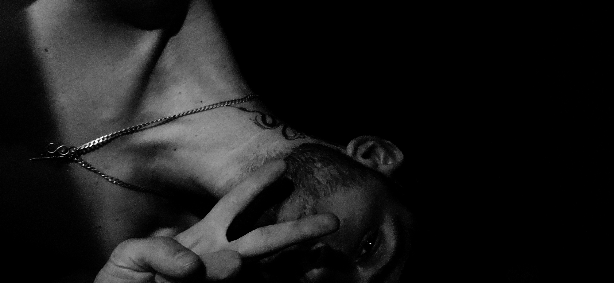 A black and white, waist-up, indoor medium shot captures a man with short hair and stubble from the chest up, angled to the right, with his head tilted down. He is wearing a silver chain with a pendant and has a tattoo on his neck. His right hand is raised, with the index and middle fingers extended in a peace sign, positioned in front of his face. The lighting is dramatic, with strong shadows obscuring much of his body and the background, which is entirely black. The man's left shoulder and upper chest are visible, along with his ear and part of his face, including his eye. The setting appears to be a dimly lit indoor space, with no discernible urban features.