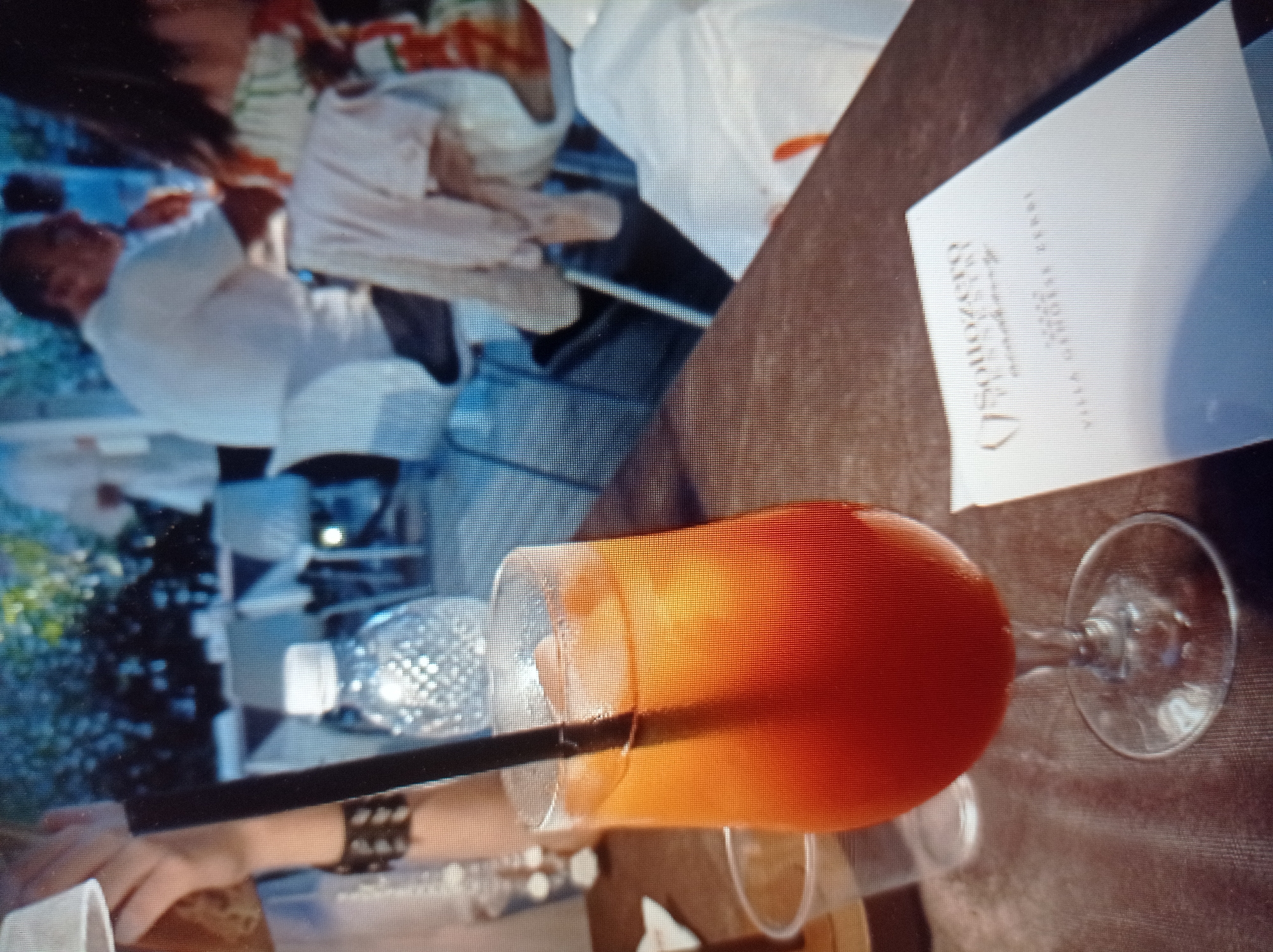 The image shows a close-up of a cocktail on a table in what appears to be an outdoor dining setting. The cocktail is a vibrant orange color, served in a tall glass with ice and a black straw. In the background, out of focus, several people are visible seated at tables, suggesting a restaurant or cafe environment. A plastic cup and a menu or flyer are also on the table in the foreground. The lighting suggests it might be daytime or early evening. There is no discernible information in the image that would indicate its specific location within the city of Mukachevo, Ukraine.