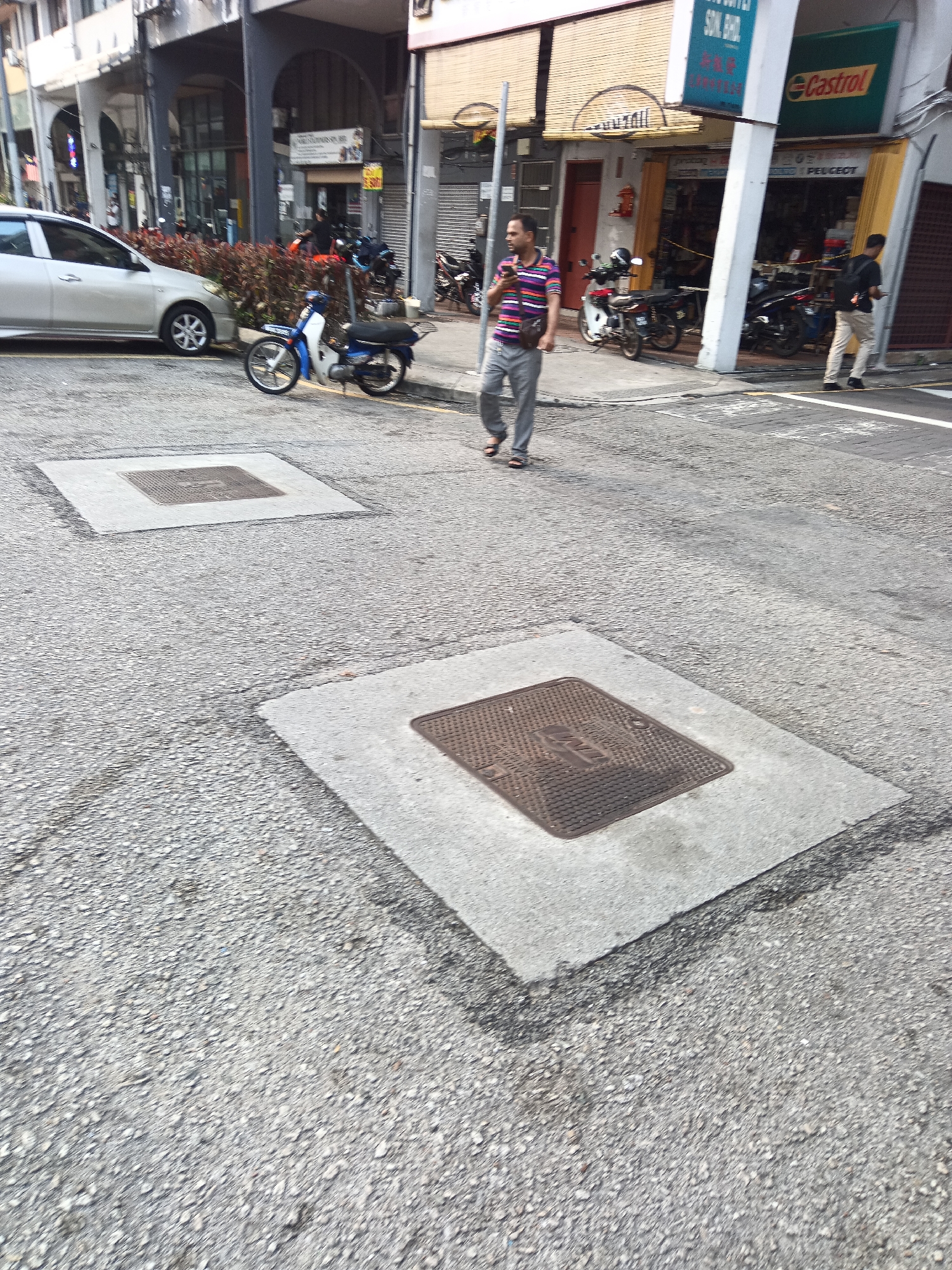 The image captures a street scene in Kuala Lumpur, Malaysia, likely in a commercial or mixed-use area. In the foreground, two concrete square slabs with metal covers are visible on the asphalt road. A man in a striped t-shirt and grey pants walks across the road, looking at his phone. To the left, a silver car is parked, and a blue and white motorcycle is also parked on the roadside. In the background, buildings with storefronts line the street, one of which displays a "Castrol" sign. Several motorcycles are parked in front of the buildings. The lighting suggests it is daytime, possibly late morning or early afternoon.