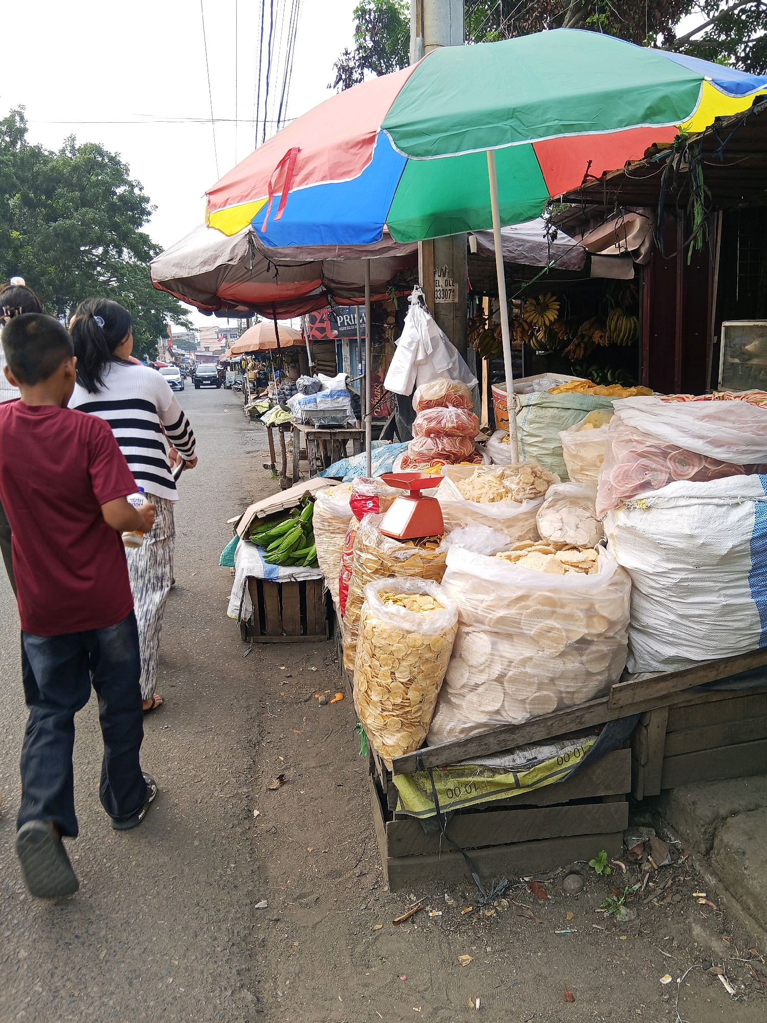 The image depicts a street scene in Lubuklinggau, Indonesia, likely a market area, with vendors displaying their goods. In the foreground, a young boy wearing a maroon t-shirt and dark pants walks away from the camera, holding an orange bottle. Behind him, a woman in a black and white striped shirt and patterned pants walks in the same direction. The street is lined with market stalls covered by colorful umbrellas, shades of red, yellow, blue, and green.
The stalls are laden with various food items, predominantly dried snacks and produce. Large transparent bags are filled with light-colored circular crackers and other fried snacks. A wooden crate contains a pile of green vegetables, possibly string beans. There are also stacks of packaged pink and red snacks. A red weighing scale is visible on one of the stalls. Bananas hang from a stall in the background.
The environment appears to be an urban street with a paved road. Trees with dense foliage are visible on the left side of the image. In the distance, cars are driving on the road, suggesting it is a functional street rather than a pedestrian-only market. Power lines crisscross the sky. The ground beside the stalls is rough and appears to be a mixture of dirt and debris, including cigarette butts. The overall impression is a bustling, open-air market on a moderately busy street.