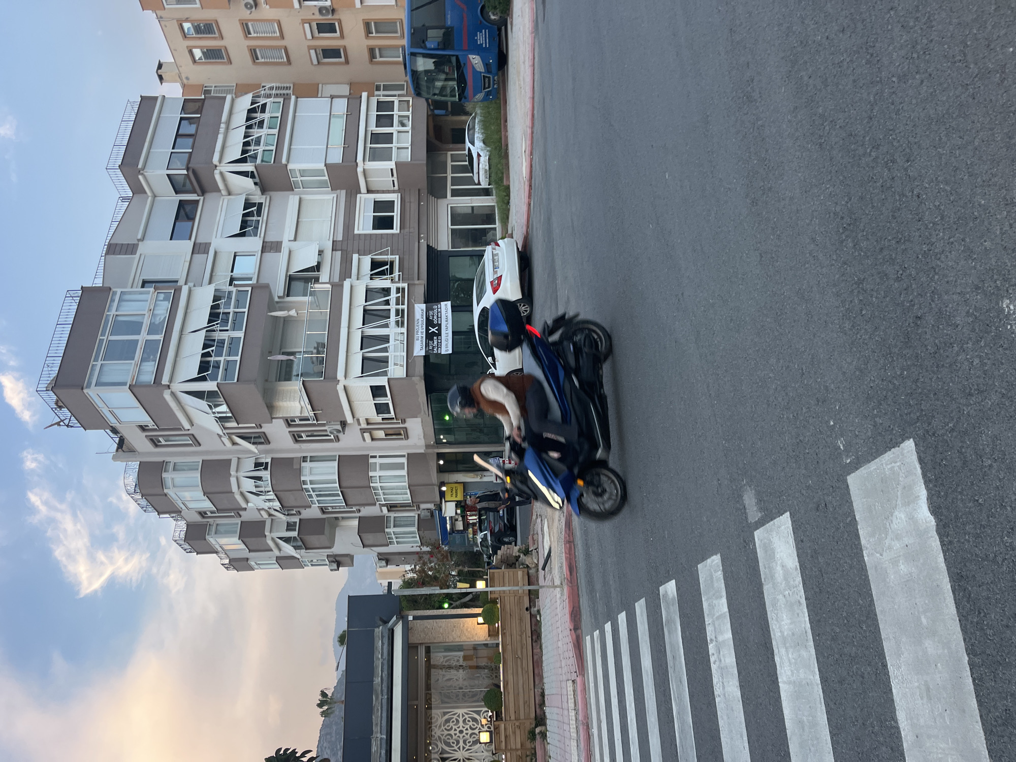 The image captures an urban street scene in Alanya, Turkey, during what appears to be either late afternoon or early morning, indicated by the soft, warm light in the sky. The perspective is slightly elevated, looking down a street lined with buildings.
In the mid-ground, a person wearing a helmet, a light-colored top, and a brown vest is riding a blue scooter or motorcycle. The scooter is on the right side of an asphalt road, approaching a zebra crossing marked by white stripes.
On the left side of the street, a prominent multi-story apartment building with numerous balconies and windows, some equipped with partially open shutters, dominates the background. Further down the road, a blue bus or van and a white car are visible. In the foreground on the left, a modern building with a decorative metal fence and illuminated exterior lighting suggests a commercial or public space. The sky above is mostly clear with a few wispy clouds, displaying a gentle gradient of blue and subtle orange hues.
On the sidewalk, near the white car, a white advertising sign is clearly visible. The text on the sign, in Turkish, reads: "YILMAZ," "BU PROJENİN TASARIM VE UYGULAMASI" (The design and application of this project), "DALOMER X GÖRGÜLÜ," "İŞ BİRLİĞİ İLE YAPILMAKTADIR" (is being done in collaboration with), and a partial phone number "4312 45 23." The scene depicts a typical moment of daily life and urban infrastructure in a Turkish city.