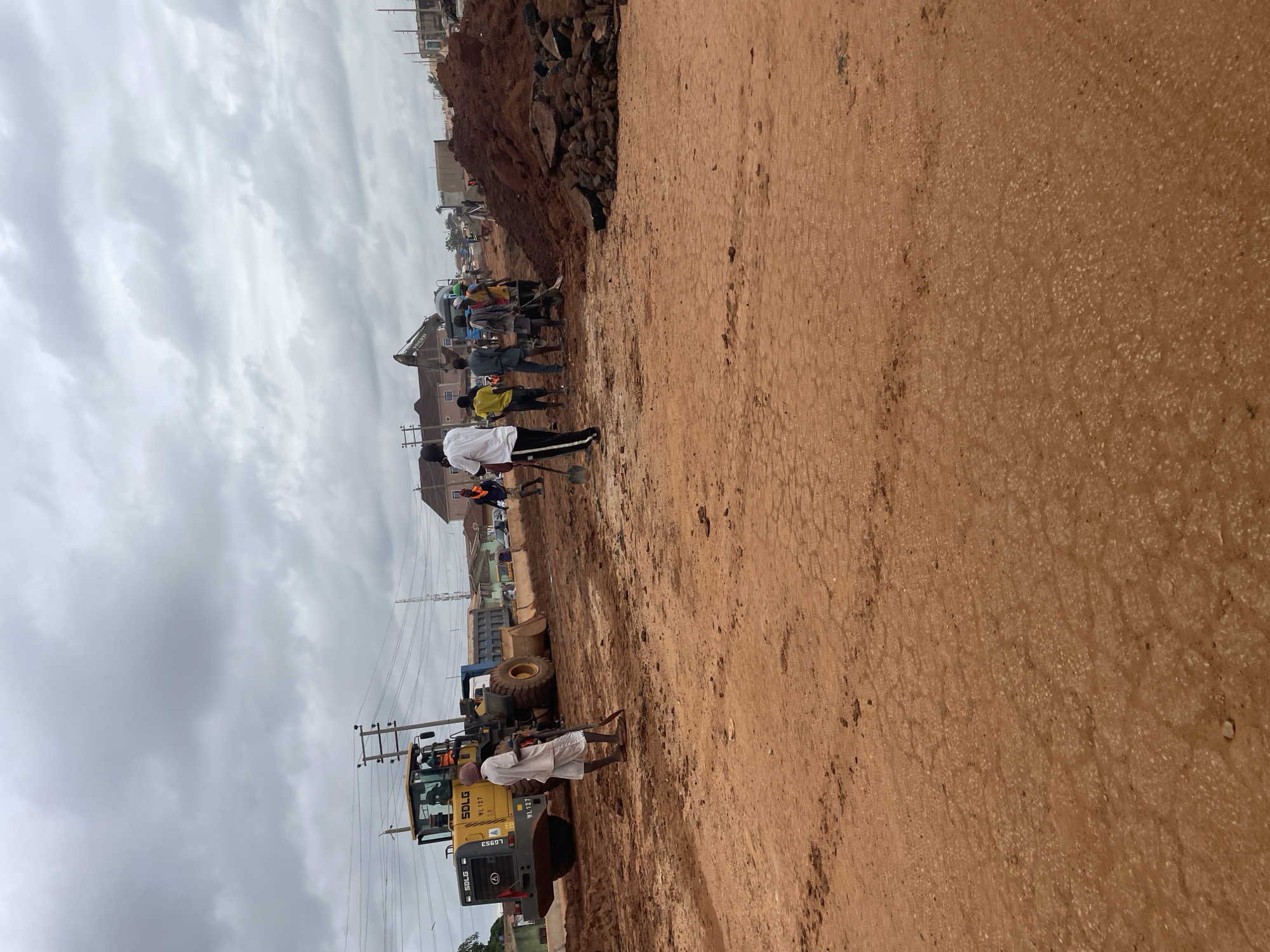 The image captures an ongoing road construction scene in Minna, Nigeria. A yellow SDLG front-end loader, model LG53, with "WL 127" visible on its side, is positioned on a dirt road. A man in a white shirt and shorts stands beside it, holding a shovel. Several other individuals, dressed in casual clothing and some wearing orange safety vests, are present, some with tools like shovels and a wheelbarrow. In the background, a Volvo excavator is visible, and a large pile of reddish-brown soil and rubble is amassed. The environment suggests a cloudy day, with power lines and buildings forming the backdrop. The ground is covered in red earth, indicating a potentially dusty or muddy construction site.