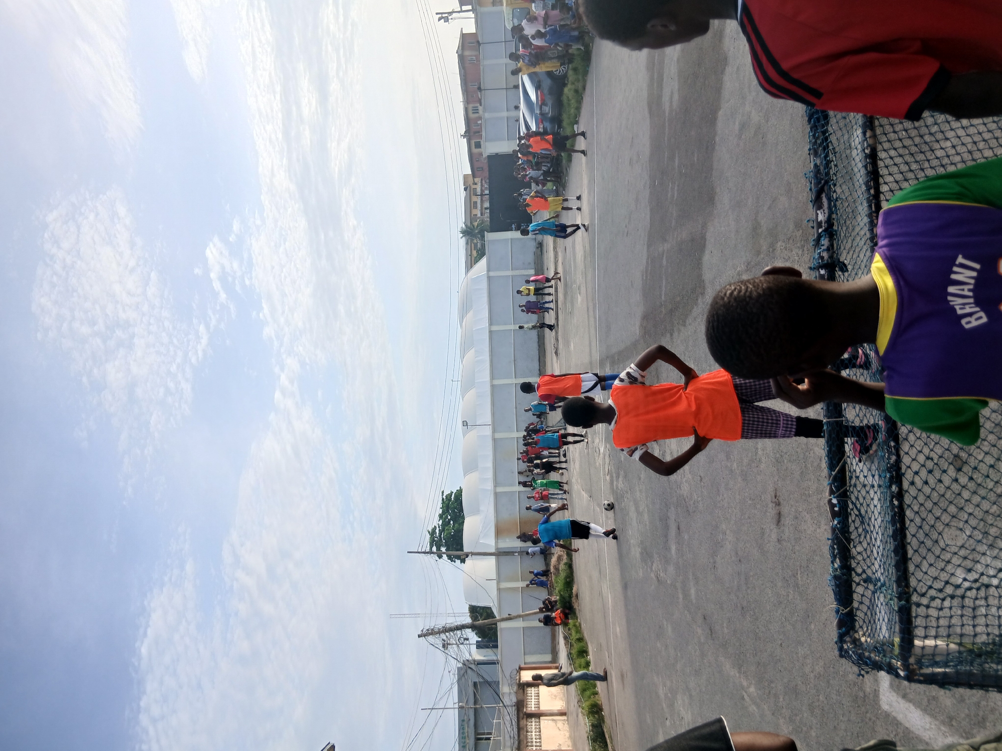 The image shows a soccer practice in Lagos, Nigeria, under a partly cloudy sky. A group of young players is practicing on an asphalt field marked with white lines. Several are wearing orange vests over their shirts. A white, arched structure is in the background, likely a covered sports facility. Buildings can be seen behind the sports facility. Power lines cross the sky. Several people are standing near a perimeter fence. The scene is observed from behind a net.