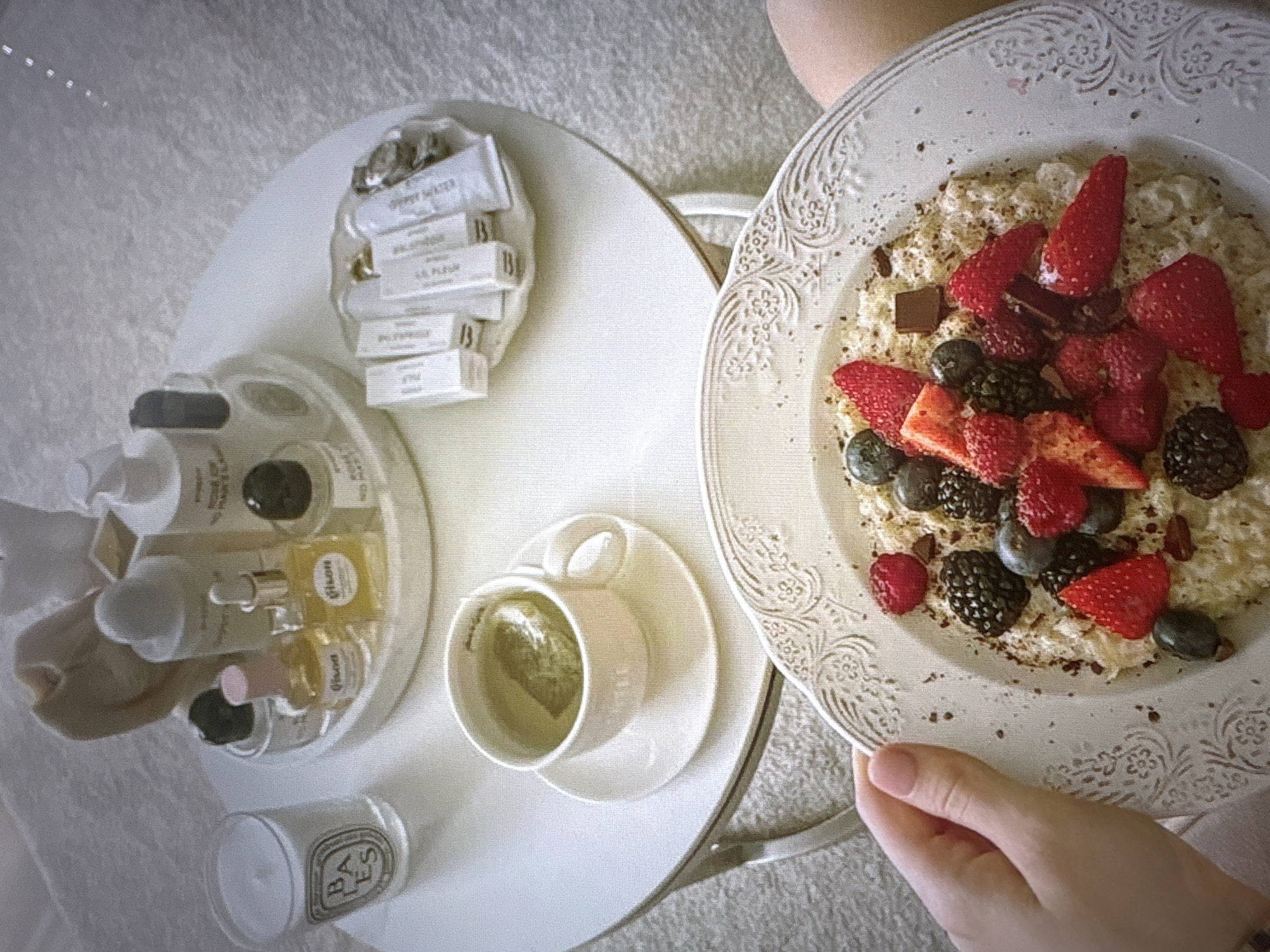 The image depicts a close-up of a bowl of oatmeal topped with various berries, including strawberries, blueberries, and blackberries, as well as a piece of dark chocolate. The oatmeal is served on a decorative white plate. In the background, a round white table holds a cup of tea, a candle in a glass holder with the "BA ES" logo, and a tray with several small bottles and tubes of cosmetics. The environment appears to be an indoor setting, possibly a bedroom or a living room, with a textured rug visible. The image is taken from a high angle, focusing on the food in the foreground. There is no information in the image to suggest the location is in Pori, Finland.