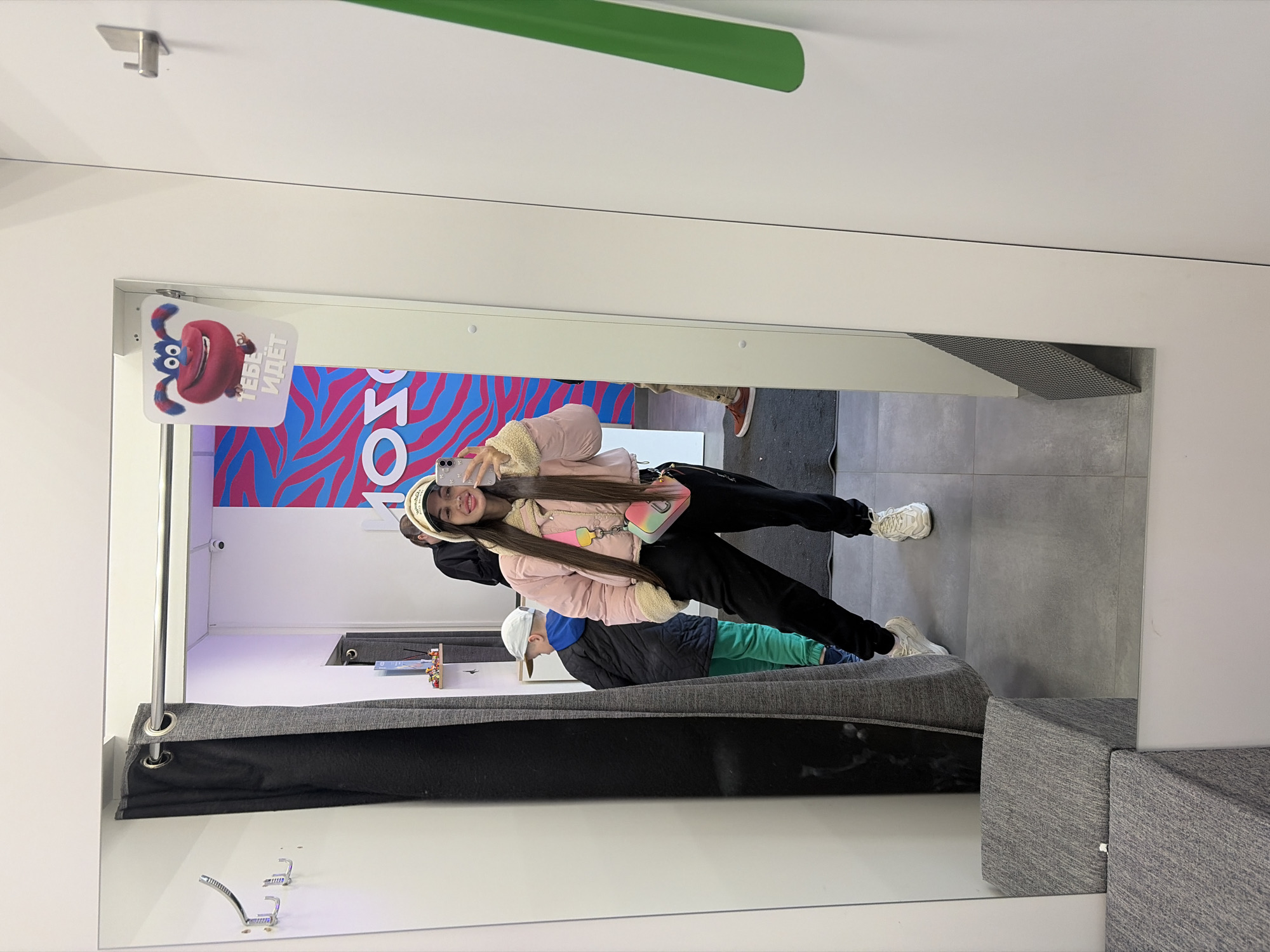 The image shows a reflection in a mirror within a fitting room. A woman in a pink jacket and black pants is taking a selfie. Behind her, partially visible, is a child wearing a white cap, a black jacket, and green pants. A gray curtain is partially drawn across the fitting room entrance. The room also contains a gray seating area. Above the mirror, a sign with a cartoon character and Russian text reading "Тебе идёт" (You look good) is visible. The reflection shows a gray tiled floor and parts of a retail store. The location is likely Saint Petersburg, Russia, based on the Russian text on the sign.