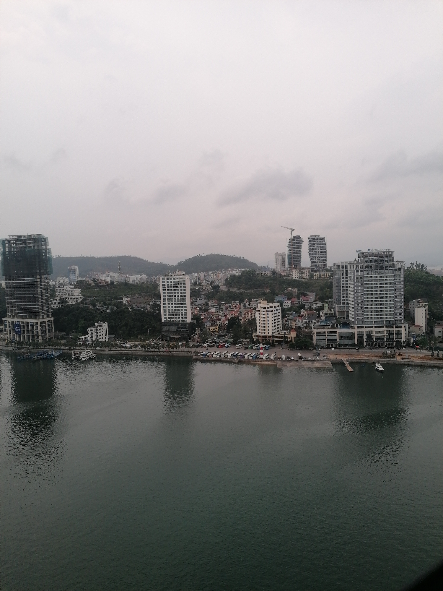 The image shows a cityscape in Bai Chay Ward, Vietnam, viewed from an elevated position overlooking the water. The sky is overcast. Several high-rise buildings are visible, including one under construction with exposed framework and netting. Other buildings are completed, with multiple stories and regular window patterns. The city is built up from the waterfront, extending onto hills in the background. A crane can be seen near the twin towers on a hill. The shoreline features a row of parked buses or vehicles, as well as smaller boats docked in the water. The water surface is calm with reflections of the buildings. The scene appears quiet and devoid of significant human activity.