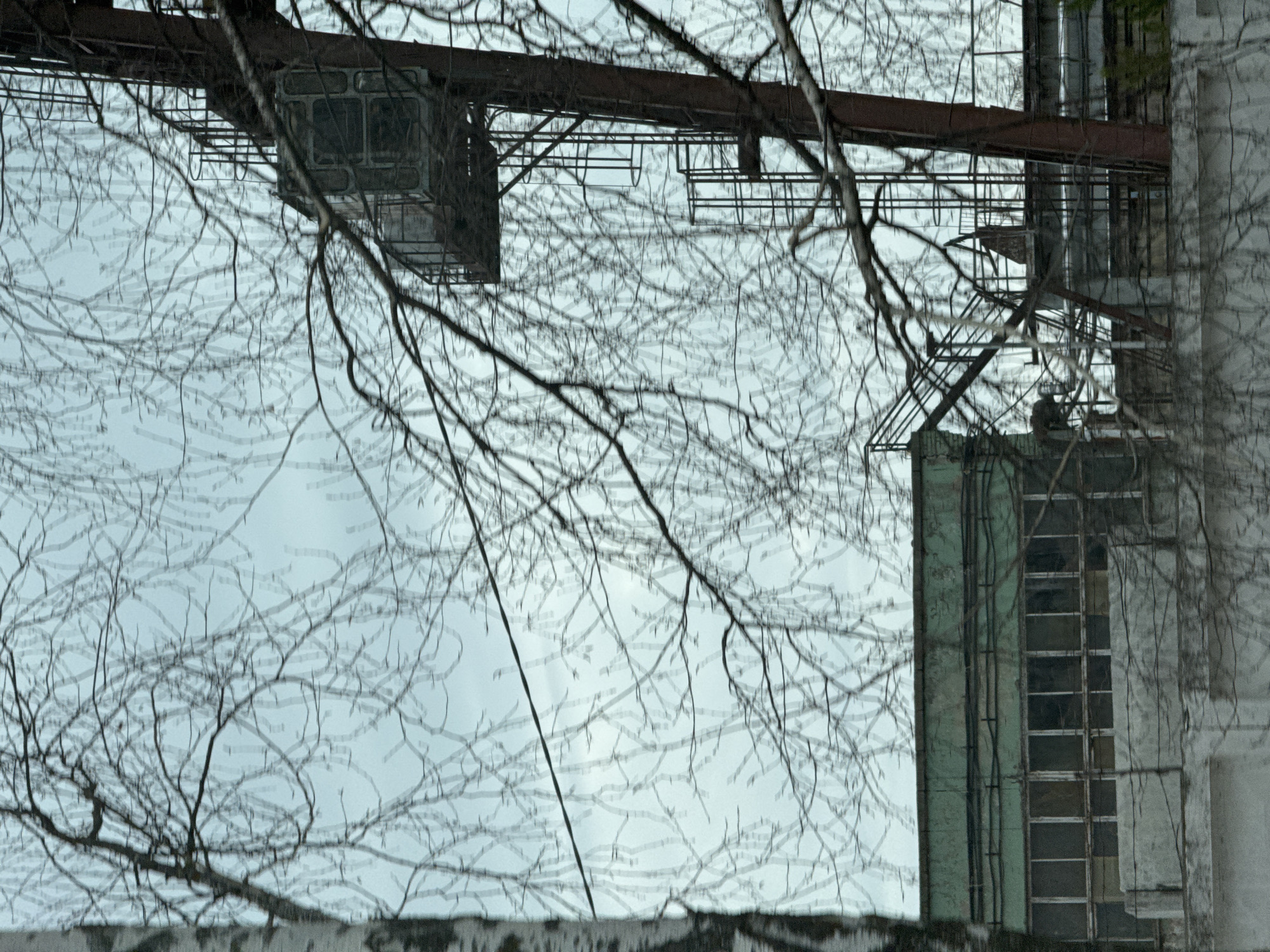 The image shows a view looking upwards, partly obscured by bare tree branches. The architecture indicates an industrial area or a structure with functional design. There are metal structures, possibly part of a crane or gantry system, overhead. A building with multiple window panes is visible on the right, its paint appears faded and weathered. The scene is likely located in the city of Chaikovsky, Russia.