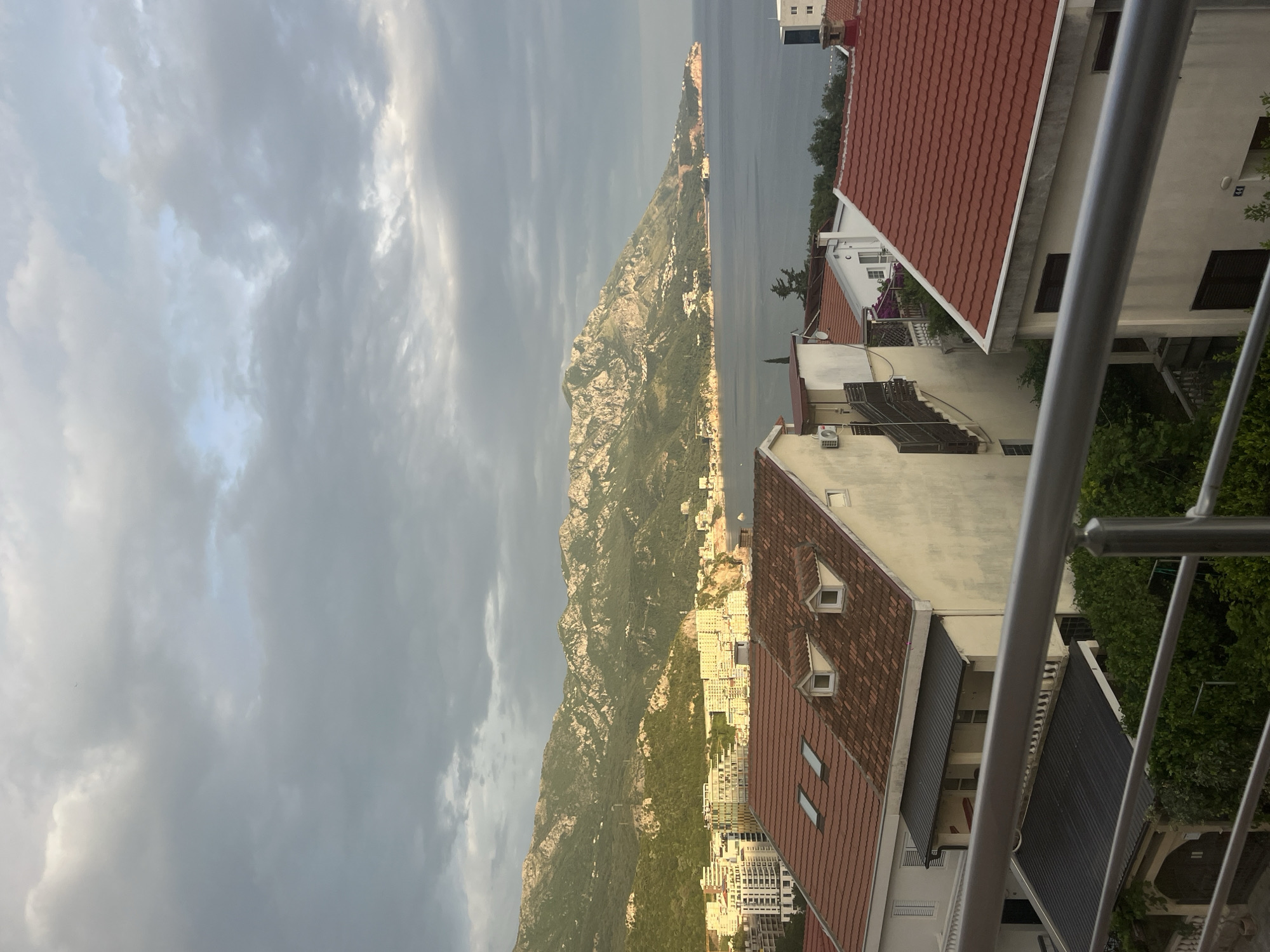 The image captures a view from what appears to be a balcony in Bečići, Montenegro. In the background, a large, rugged mountain rises dramatically against a cloudy sky. The mountain runs vertically down the left side of the frame.
To the right of the mountain and between the mountain and the water, the cityscape of Bečići is visible, consisting of buildings of varying heights with predominantly light-colored facades and red-tiled roofs.
In the foreground, building rooftops, including red-tiled roofs, are visible. Metal balcony railings run vertically. The sky is overcast.