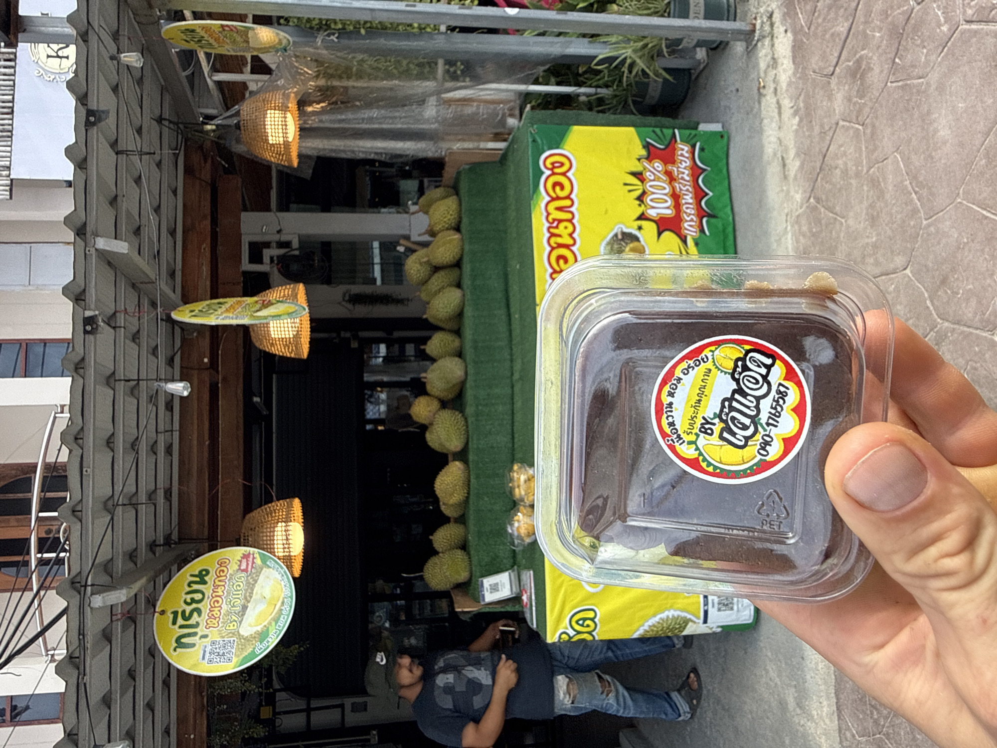 A person is holding a small, clear plastic container with a dark substance inside, labeled with a colorful sticker in Thai and a ♷ PET recycle symbol. In the background, there's a shop selling durian fruit. The storefront is visible with durians stacked on a green-topped display. Signs are present, also in Thai, advertising the durian. A man, wearing a t-shirt, jeans, and a hat, is standing near the display. The scene is shot in daylight, with a view of the street and building exterior. The sign at the street is a durian vendor in Moo 1, Thailand.