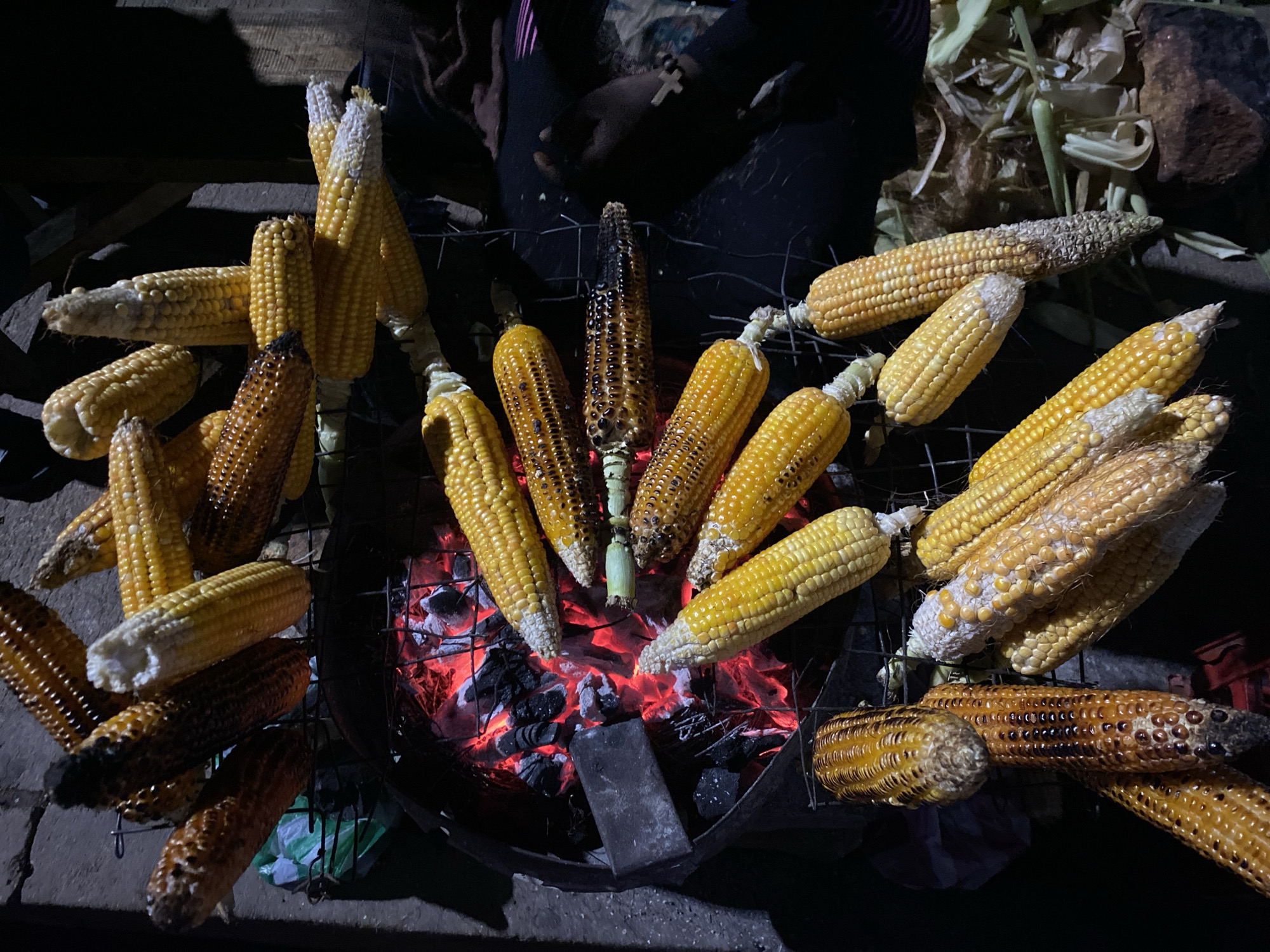 A top-down shot shows grilled corn on the cob over burning coals. The corn is arranged on a metal grill, with some cobs still yellow and others browned or charred from the heat. In the background, a person sits near the grill. To the right, some corn is piled next to the grill with the husk still attached. This photo was taken in the city of Bukuru, Nigeria.