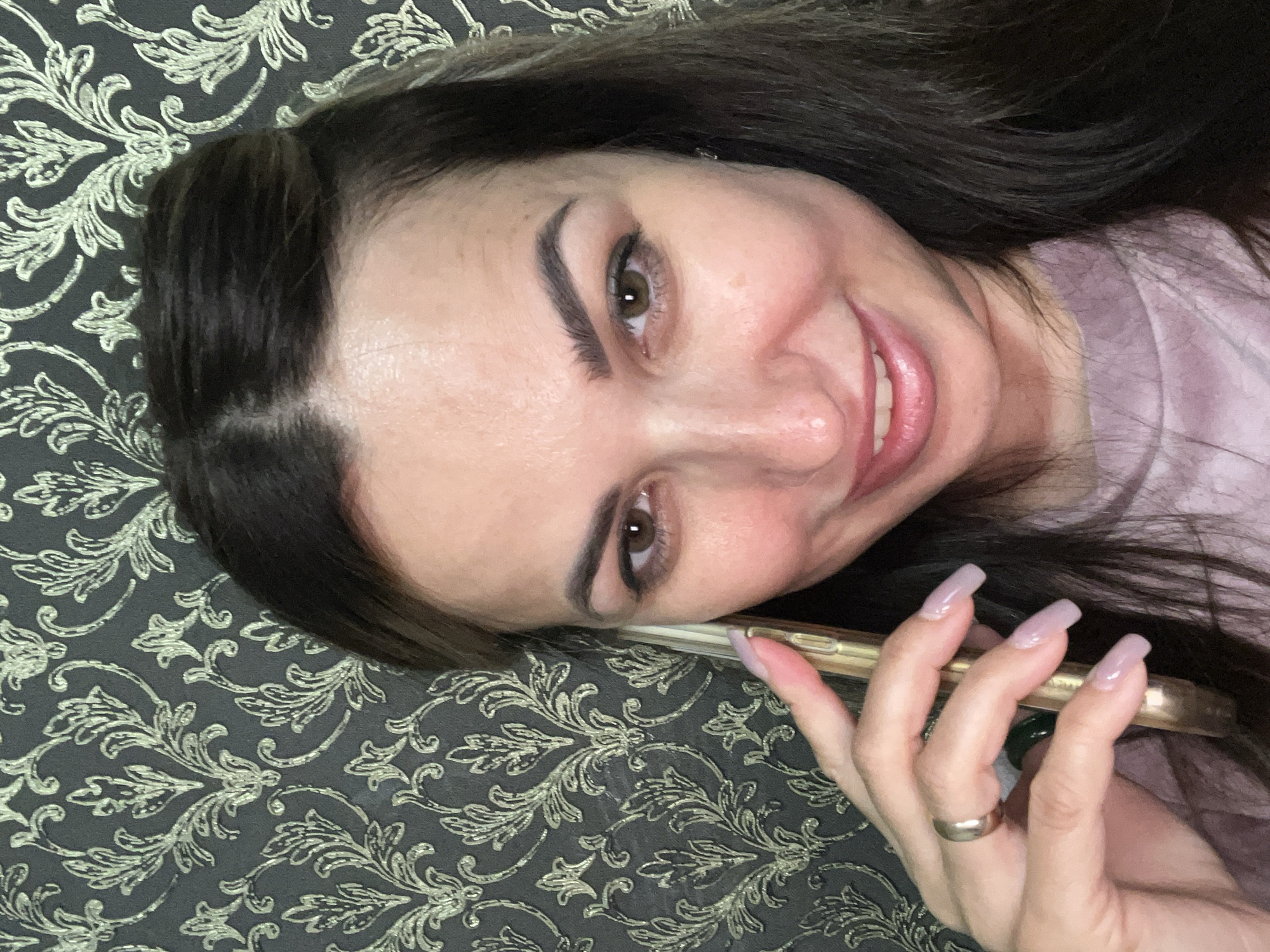 A woman with dark hair is holding a gold-colored smartphone to her right ear, smiling at the camera. She is wearing a light purple, crew-neck shirt. A ring is visible on her left ring finger. The background is a dark wallpaper with a gold floral pattern.