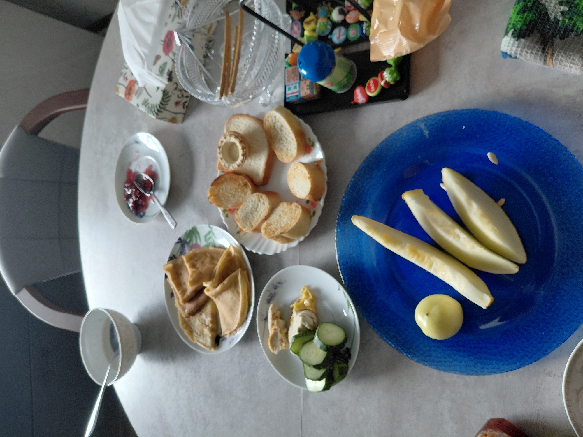 The image shows a table with various food items, including pancakes, bread, sliced cucumbers, fruit, and a bowl of liquid. There are also some decorative items and a napkin. The scene appears to be set indoors, possibly in a dining area or kitchen. The specific location in Novosibirsk, Russia cannot be determined from the image alone.