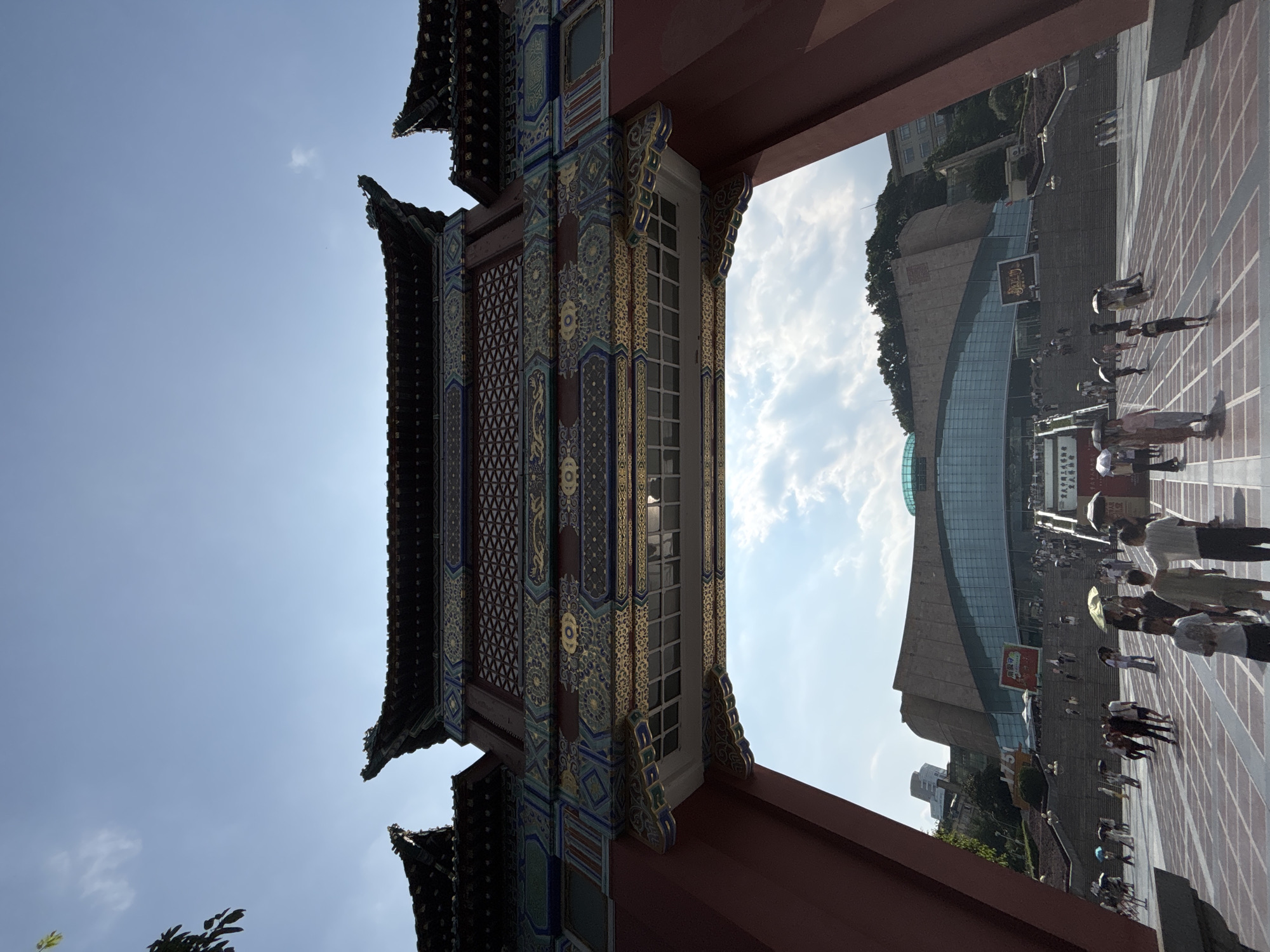 An exterior, low-angle, wide shot captures a traditional Chinese gate structure framing a modern museum. The gate is elaborately decorated with red paint, intricate gold, blue, and green patterned carvings, and curved eaves with dragon-like embellishments. The sky above is bright blue with scattered white clouds.
Through the archway of the gate, a large, contemporary museum building is visible. The museum features a prominent glass facade and a sloping, stone-clad upper section. Several tiered staircases lead up to the museum entrance, where a crowd of people can be seen ascending. Many individuals are holding umbrellas, suggesting it is sunny or there is a possibility of rain.
The ground in front of the gate and leading to the museum steps is a wide, open plaza paved with a repeating square tile pattern. People are scattered across the plaza, some walking, some standing, creating a sense of activity and public gathering. The location is identified as Yuzhong District, China.