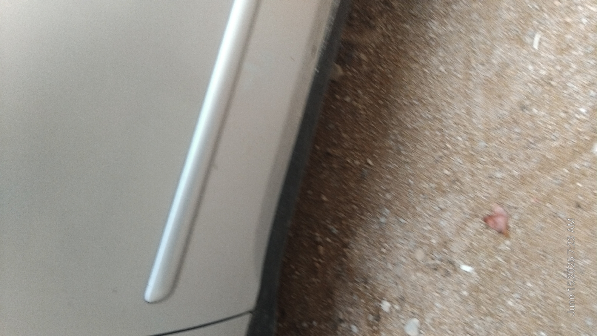 The image displays a close-up, low-angle view of the lower side panel of a light-colored vehicle, likely a car, situated on a granular ground surface. A prominent horizontal trim piece, appearing metallic or silver, runs along the car's body. The ground is composed of small pebbles and sand, with some debris scattered across it, including what appears to be a small, reddish-brown leaf in the lower central portion of the frame. The bottom right corner of the image shows a timestamp: "June 15, 2026 7:23 AM". There are no people or discernible activities visible. The scene suggests an outdoor setting, possibly a street or parking area, but offers no specific indicators of the city of Howayi, Nigeria.