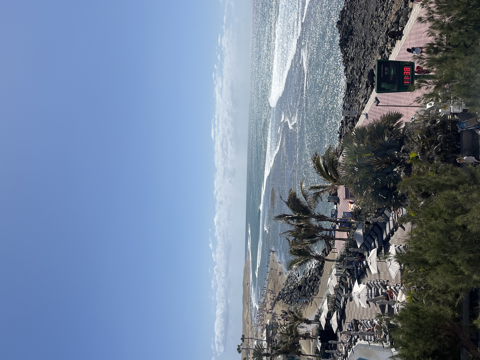 The image shows a beach scene in Maspalomas, Spain. The view is from an elevated position, looking down towards the beach and ocean. The sky is blue with some clouds. The ocean has waves. A sandy beach is visible with people walking and sunbathing. There are palm trees and other vegetation. A promenade runs along the edge of the beach with people walking on it. Lounge chairs with umbrellas are on the beach. A digital display shows the time as 11:38.