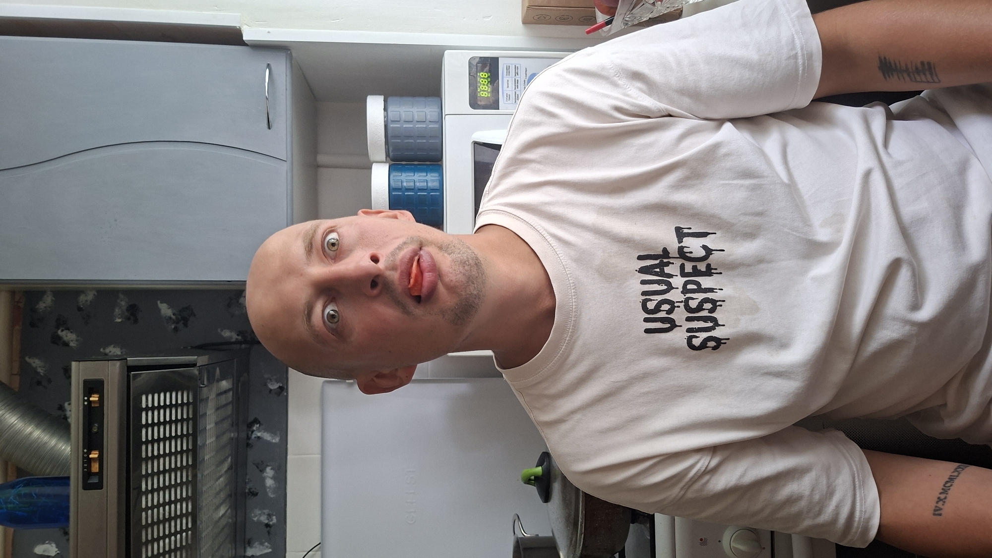 A bald man with light-colored eyes and a goatee is pictured from the chest up, in what appears to be a domestic kitchen. He is wearing a cream-colored t-shirt with the words "USUAL SUSPECT" printed in black, dripping letters on the front. The man has a piece of red food, possibly a tomato, held between his teeth and tongue, with his eyes wide open and looking directly at the camera.
The background shows various kitchen elements. To the left, a stainless steel range hood is visible above a stovetop with pots. A white refrigerator is to the far left. Behind the man, on a shelf above a microwave, are two blue and gray cylindrical containers. The microwave itself displays a digital time of "88:88". The walls have a grey, patterned wallpaper.
On the man's left arm, a tattoo with Roman numerals, "IV.X.MCMLXXXI", is visible. On his right arm, another tattoo of thin, vertical lines resembling a sound wave or barcode is present. The overall lighting suggests it is daytime, with light coming from the right side of the frame. The environment appears to be a typical kitchen setting. The specific location in Myadzyel, Belarus cannot be determined from the visual information provided.