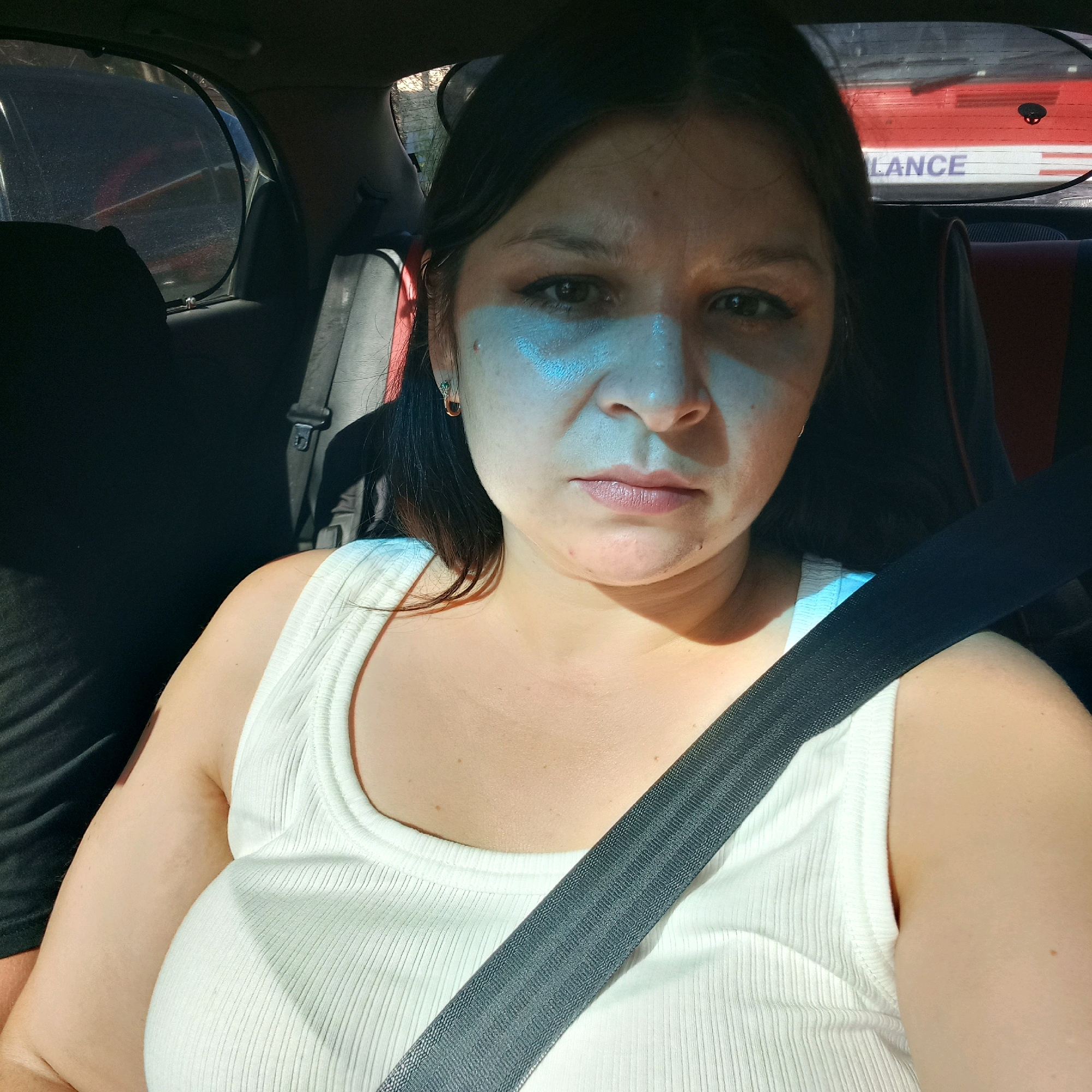 A woman is in the passenger seat of a car, looking directly at the camera. She has dark hair, brown eyes, and is wearing a white ribbed tank top. A seatbelt is visible across her chest. The background shows the interior of a car, with a window on the left side revealing trees and a glimpse of another vehicle. Through the rear window, text on another vehicle is partially visible, reading "LANCE". The scene appears to be outdoors during the daytime. No specific location within Kropyvnytskyi, Ukraine, can be determined from the image.