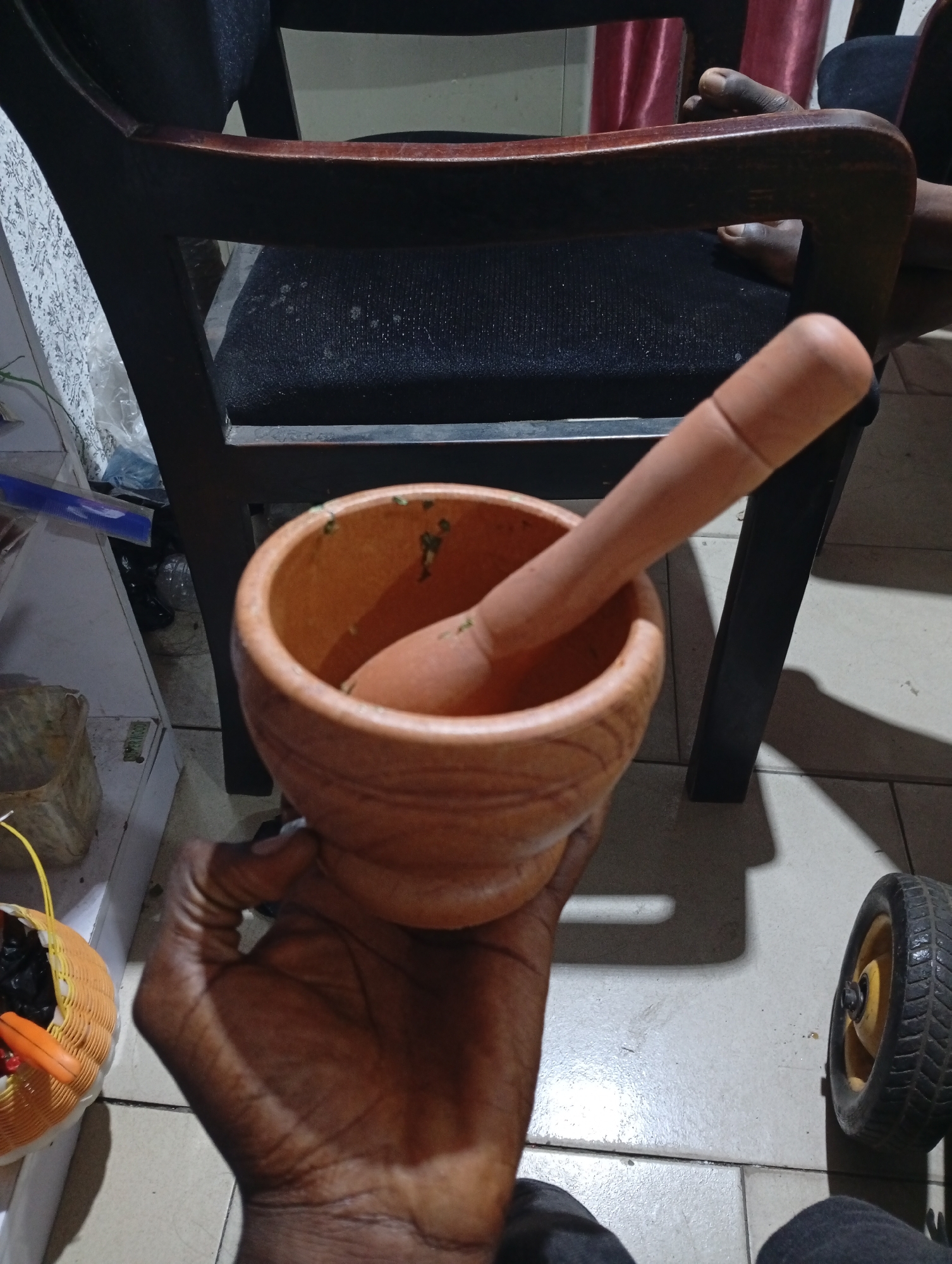 A dark-skinned hand holds a clay mortar and pestle containing green herbs. The scene is illuminated by artificial light. The mortar and pestle are textured with ridges. The pestle sits inside the mortar. A portion of a black wooden chair is visible, with a black cushioned seat. Part of a white-painted shelf with various items, including a small orange basket with black contents, is visible on the left. A small wheel is present on the lower right. The setting is likely indoors, on a tiled floor. The location within Lokoja, Nigeria, cannot be determined from the image.