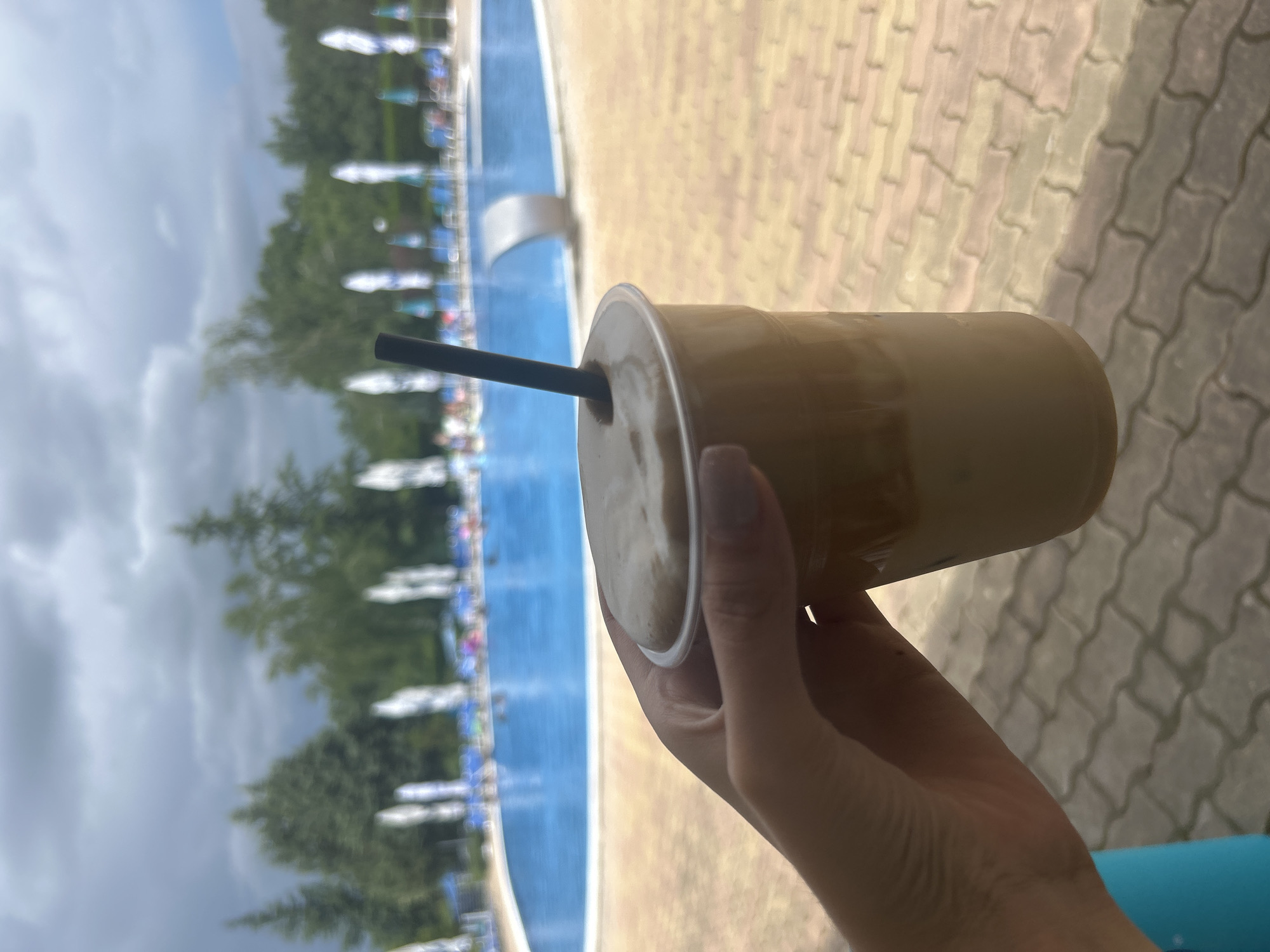 An over-the-shoulder shot captures a hand holding a plastic cup with a straw in it, filled with a creamy, light brown beverage, likely a blended drink or coffee. The cup is positioned in the foreground, slightly to the right of center. In the background, a large blue swimming pool is visible, with lounge chairs and umbrellas set up around its perimeter. Beyond the pool, lush green trees form a dense backdrop, and a cloudy sky with dark, dramatic clouds looms overhead. The ground in the foreground is paved with tan and brown interlocking bricks. The overall scene suggests a relaxing day at an outdoor pool.