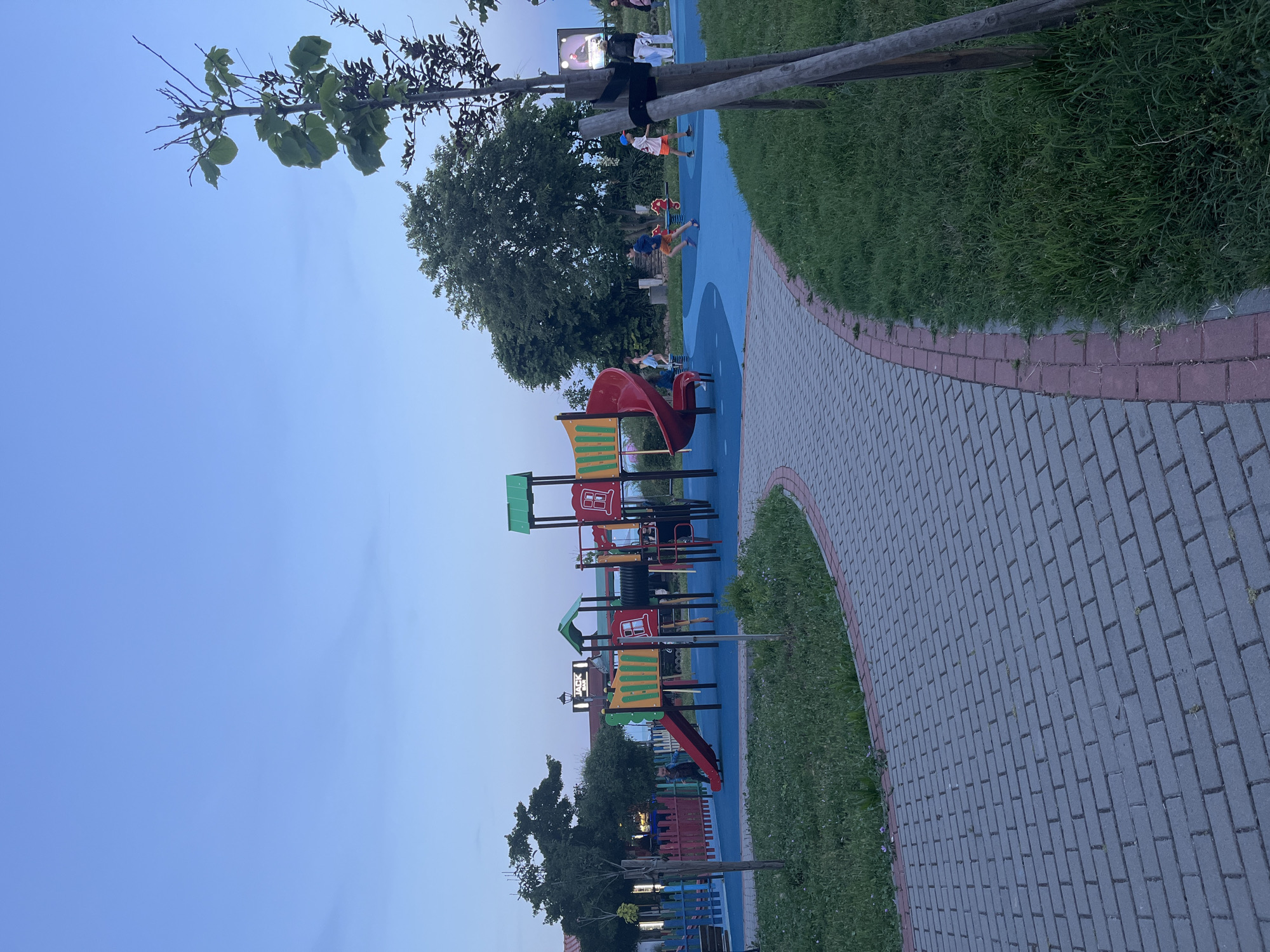 This is an outdoor photograph taken at dusk in a children's playground located in Sozopol, Bulgaria. The playground has a blue rubberized surface with a lighter blue winding path. Dominating the scene is a colorful, multi-level playground structure featuring red and yellow slides, a green roof, and red climbing bars. A group of children are visible playing on and around the playground equipment. To the left of the playground, a pink and blue fence encloses a grassy area with a small tree. A paved walkway with interlocking gray bricks and a red border curves through the foreground of the image. In the background, trees and buildings can be seen, including a sign that reads "JACK BAR." The sky is a clear, pale blue, indicating twilight.