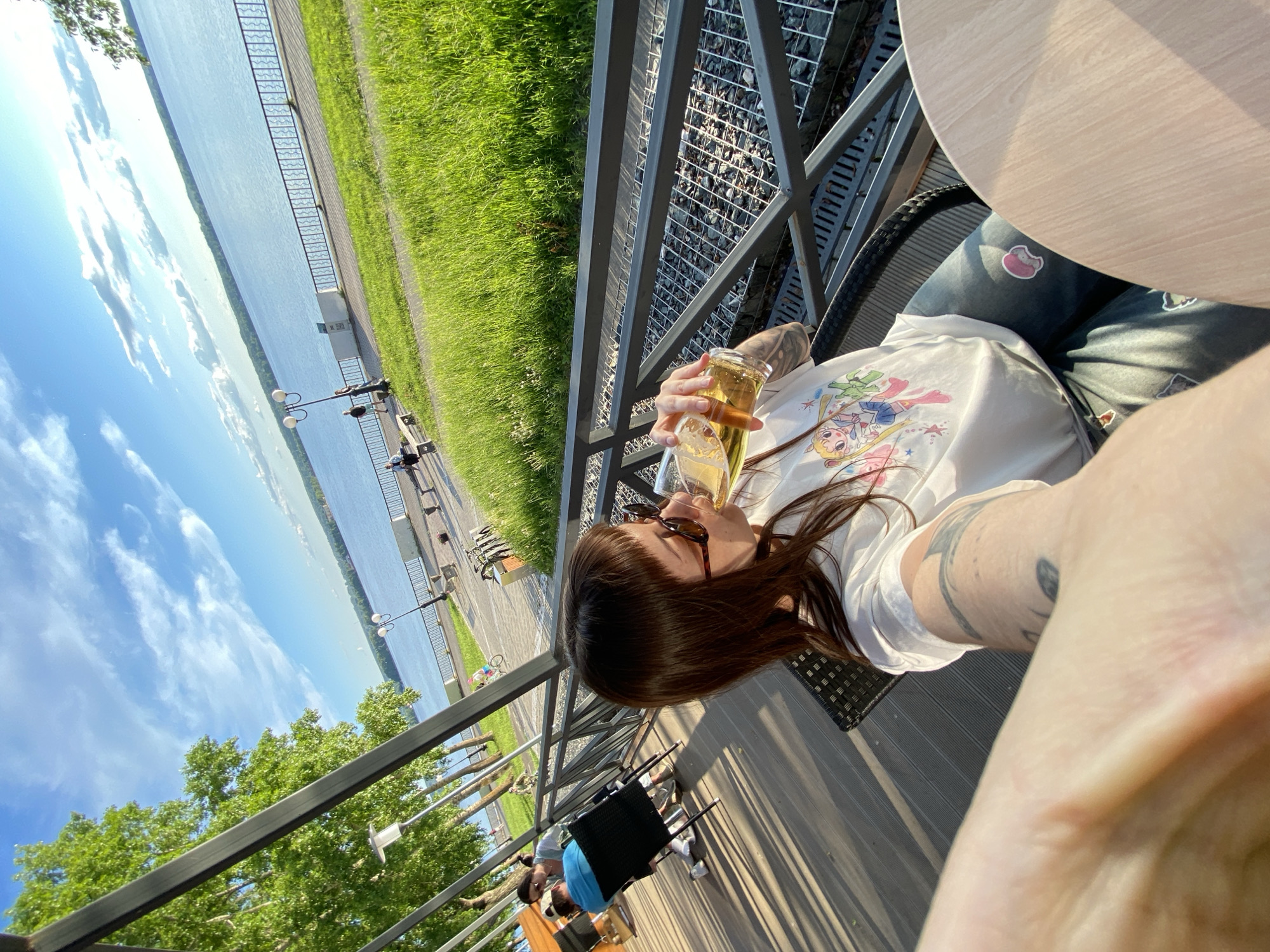 The image presents a first-person perspective from an outdoor cafe or restaurant terrace in Votkinsk, Russia, offering a scenic view of a wide body of water, likely the Votkinsk Reservoir. The day is bright and sunny, with a clear blue sky dotted with wispy white clouds, suggesting pleasant weather in the late afternoon or early evening due to the long shadows.
In the immediate foreground, a person is seated, holding a glass filled with an amber, foamy drink, possibly beer. The individual is wearing a white t-shirt adorned with a colorful anime-style print, reminiscent of Sailor Moon, and blue jeans featuring small, whimsical stickers, including one that appears to be a pink pig or cat. Sunglasses are worn, and tattoos are visible on the person's arm. Parts of a light-colored wooden table and a dark, wicker-style chair are also visible.
Beyond the metal railings of the terrace, a picturesque waterfront unfolds. A vibrant green grassy embankment slopes down to a paved walkway that runs alongside the calm, expansive water. Several individuals are present in the background, some strolling along the path while others are seated on benches. Streetlights line the walkway, and lush green trees are visible, particularly on the distant bank of the water, contributing to the serene atmosphere.
The scene captures a moment of relaxation and leisure, with people enjoying the sunny weather and the scenic waterfront view. No explicit readable text is visible, apart from the design on the t-shirt and the decorative stickers on the jeans.