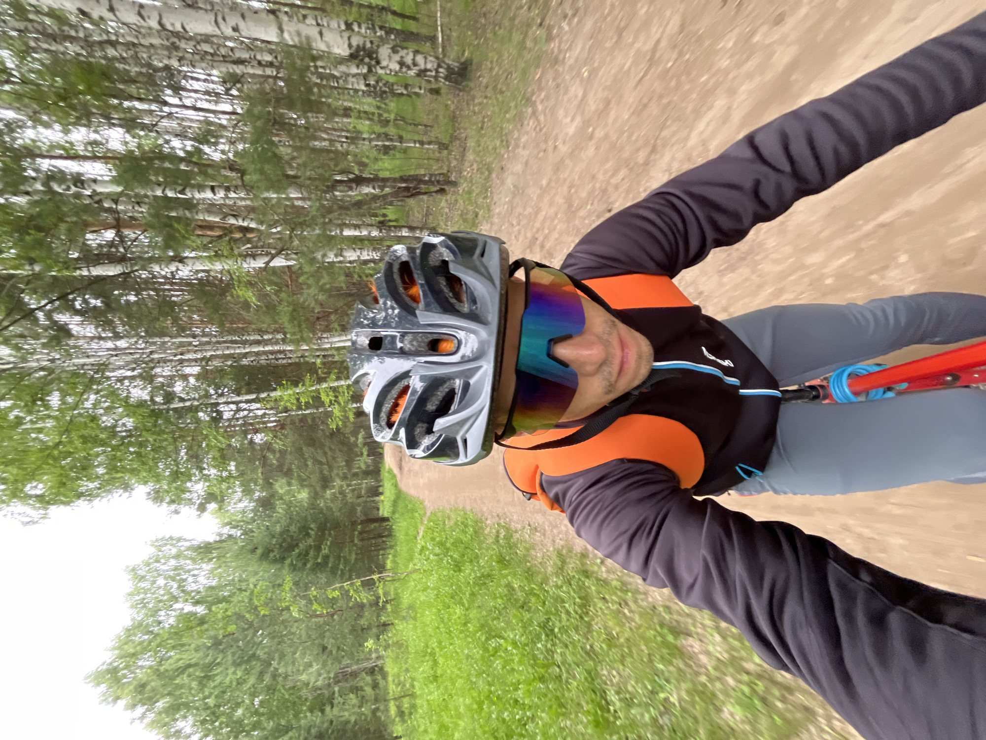 A first-person perspective shot shows a male mountain biker on a dirt path in a forest with birch and pine trees. The biker is wearing a gray helmet with orange accents and reflective, multi-colored cycling glasses. He has on a black long-sleeved jersey with an orange backpack. The path ahead is a light brown dirt track winding through a wooded area with green grass on the left side. The sky is overcast and white. This appears to be a trail in the forests near Chaikovsky, Russia.