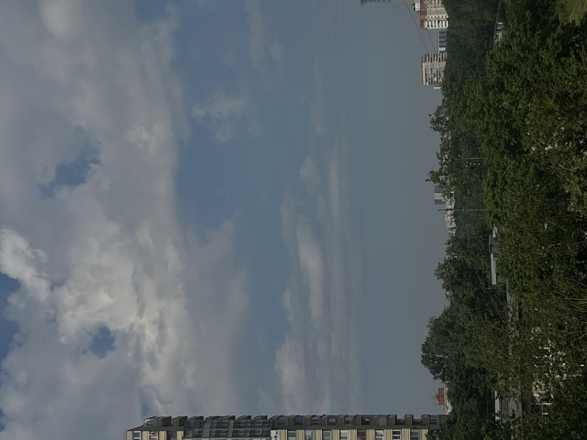 The image captures an urban landscape with a prominent tall apartment building on the left side, characterized by its yellow facade and multiple balconies. The sky above is partially obscured by large, white, and gray clouds, with patches of blue sky visible. In the middle ground, a dense line of green trees stretches across the scene, forming a natural barrier between the foreground and the distant cityscape. Beyond the trees, several multi-story buildings, including apartment complexes and industrial structures, are visible. The overall impression is that of a moderately cloudy day in a developed urban area, with residential buildings dominating the scene.