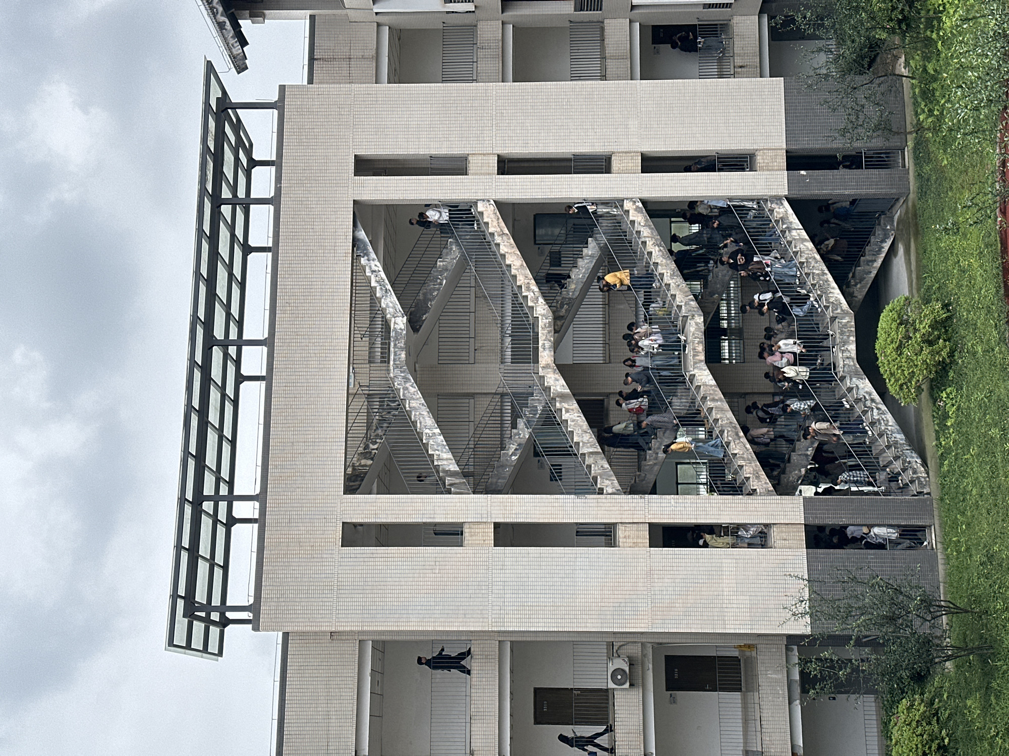 A multistory building with external staircases is shown at a tilted angle, revealing numerous individuals using the stairs and residing in dorms with the windows open. Trees and greenery line the right side of the building, and a cloudy sky is in the background. The location is presumed to be in Jiangjin, China.