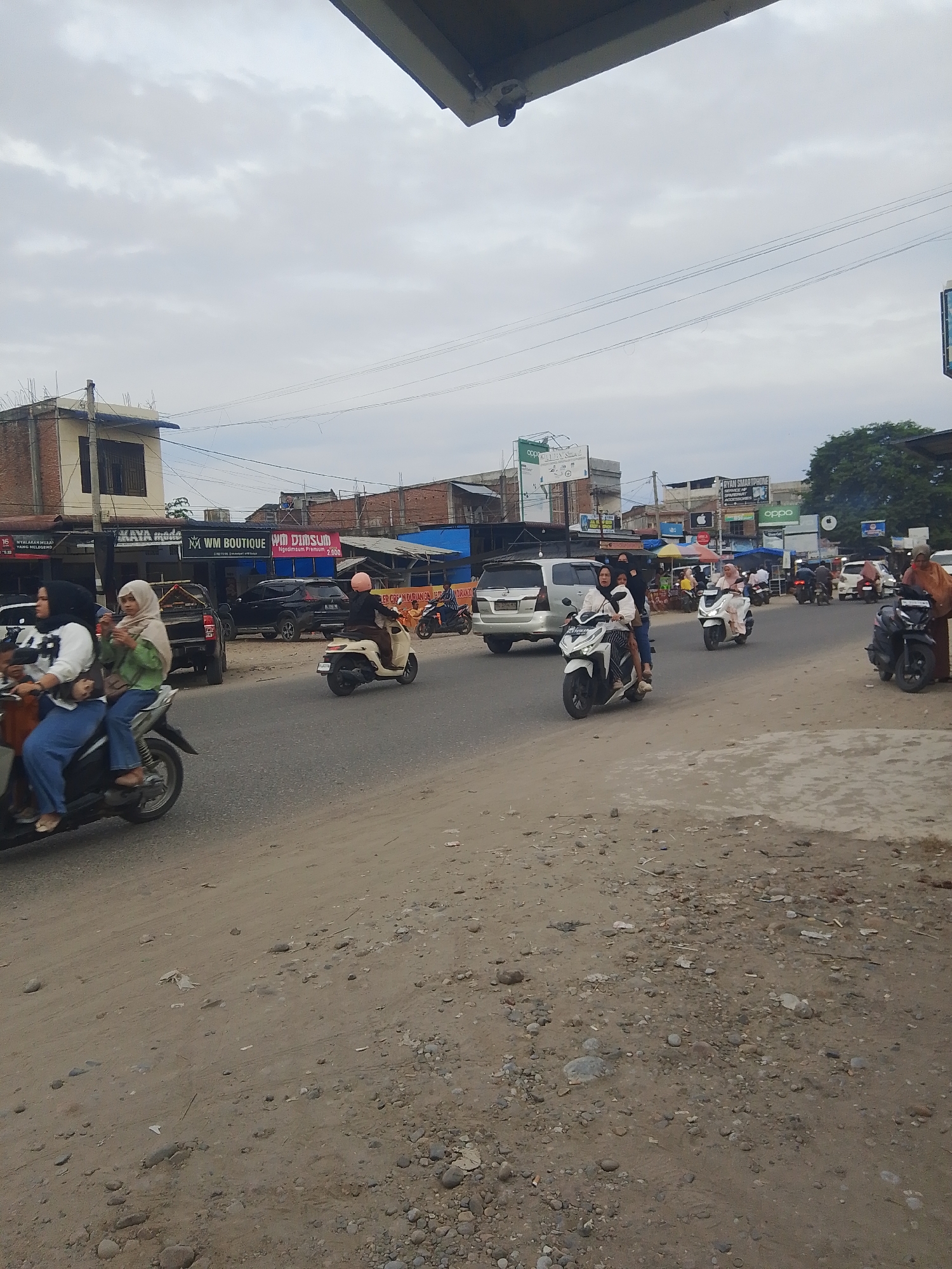 The image depicts a street scene in Gampongpaya, Indonesia, under an overcast sky. The foreground shows a dirt road with scattered rocks and debris. A grey asphalt road is to the right, carrying traffic. Multiple individuals on motorcycles are visible. In the left foreground, two women are riding a white scooter; the woman in front is wearing a white top and blue pants, while the woman behind her wears a green hijab and top. Further down the road, a woman in a white hijab and top is riding a white scooter, and another woman in a brown hijab and dress is standing next to a dark motorcycle on the right side of the road. Several cars and other motorcycles are also present in the midground and background, suggesting a moderately busy urban environment. Buildings line the side of the road, some with shops displaying signage. One sign reads "KAYA Mode" and another "WM BOUTIQUE". Another sign indicates "RYAN SMARTPHONE". The overall atmosphere is that of a typical day in a developing urban area with significant motorcycle traffic.