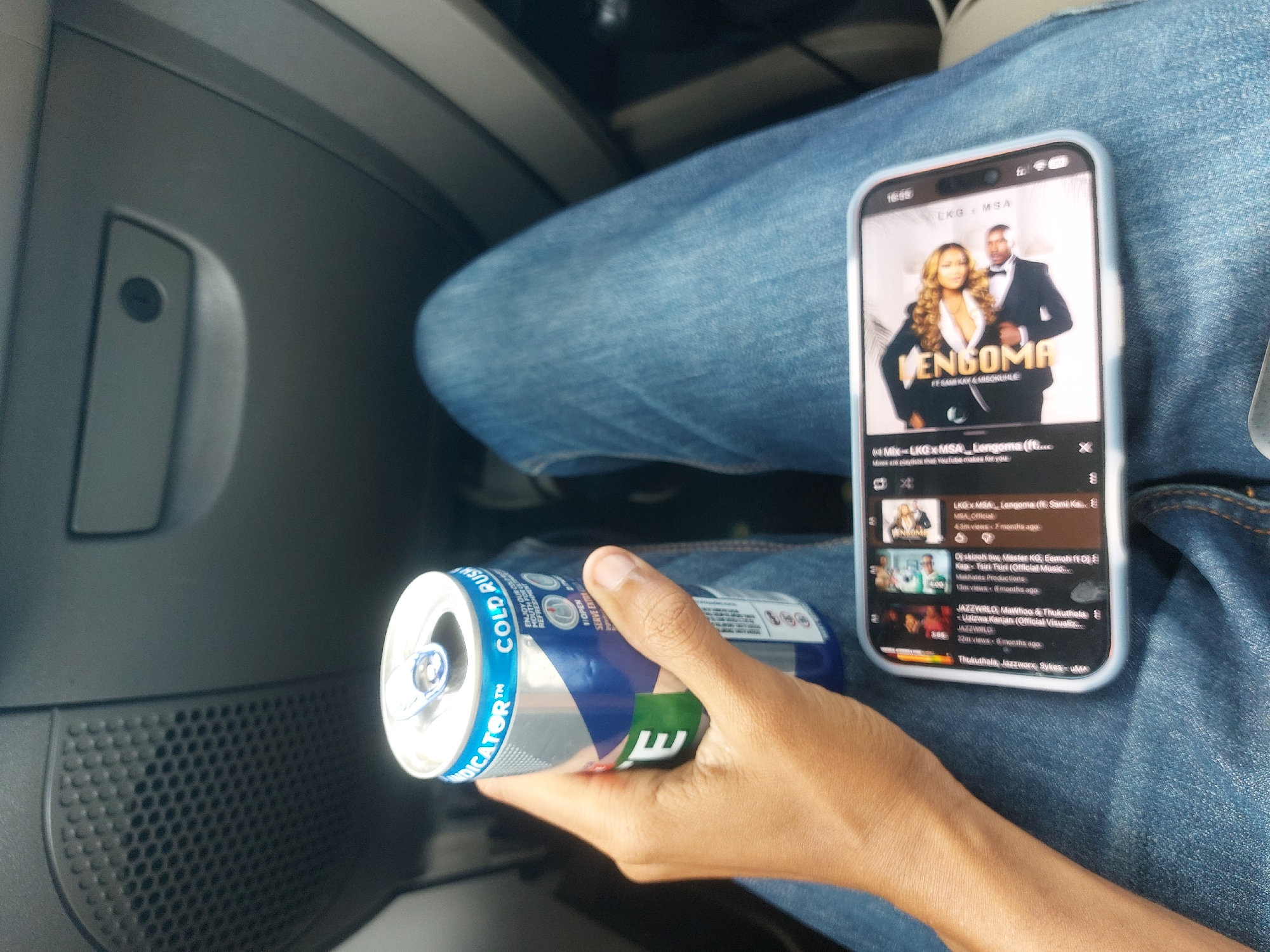 A person sits inside a car, holding a can of "Cold Rush Indicator" energy drink in their left hand. The can is blue and silver with green accents. The person's right leg is visible, clad in blue jeans. On their leg rests a smartphone displaying a YouTube playlist. The top video is titled "LKG x MSA - Lengoma ft. Sami Kay & Misokuhle." The phone shows the time as 16:59. A portion of the car's interior is visible, appearing grey with a built-in speaker. The location is likely Dar es-Salaam, Tanzania, inferred from the Swahili language used in the song title and the presence of South African artists. The lighting suggests it is daytime.