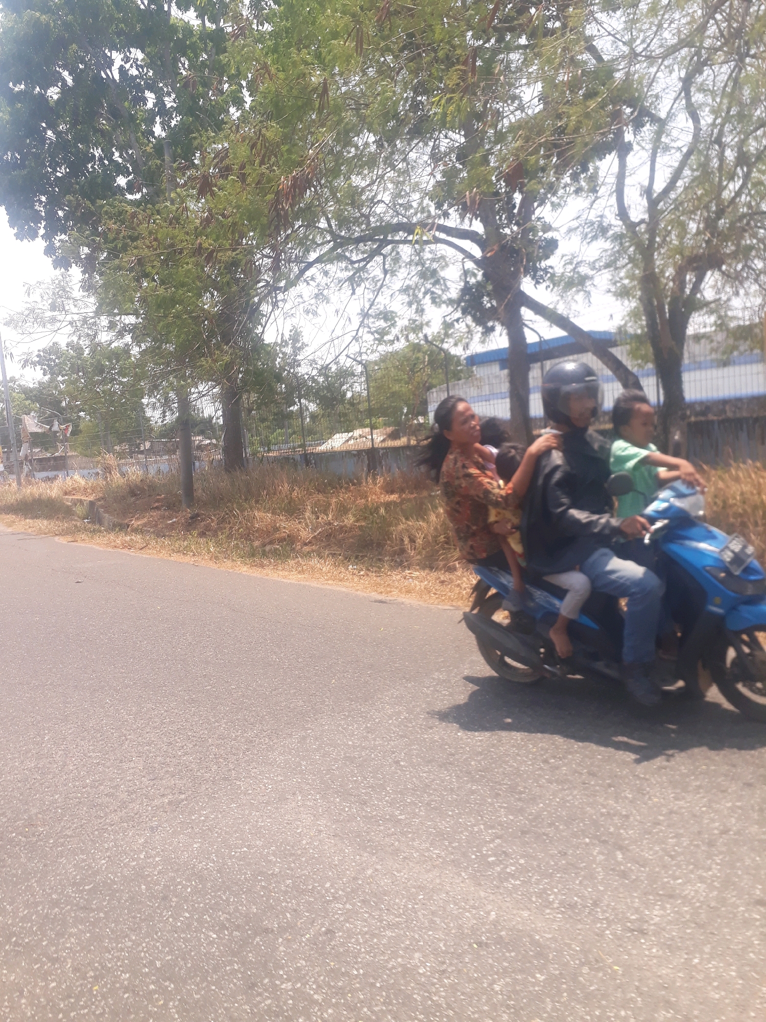 A group of four people, including two children, are riding on a blue scooter on a paved road. The driver, a man wearing a black jacket and helmet, is in the front. A woman in a floral shirt is sitting behind him, holding a young child. Another child in a green shirt is sitting on the back of the scooter. The scene appears to be in a tropical climate, with lush green trees and dry grass lining the road. In the background, a fence and some buildings are visible. The overall impression is that of a busy street scene in a Southeast Asian city.