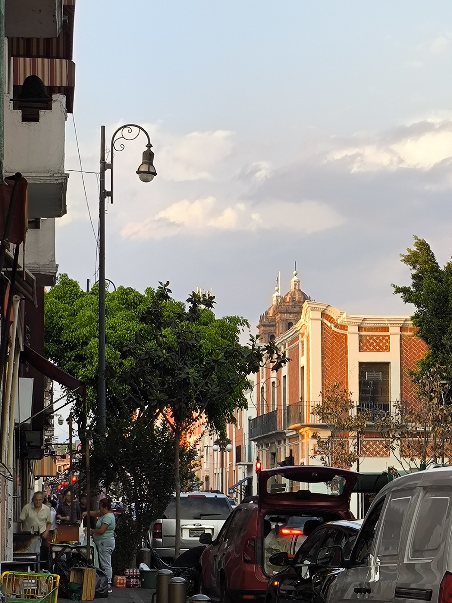 The image depicts a street scene in Puebla, Mexico, at what appears to be dusk.
**Environment:** The street is lined with buildings of varying heights and architectural styles. In the background, a prominent church with two domes is visible. Trees punctuate the scene, offering patches of green amidst the urban landscape. The sky is a mix of blue and soft, muted clouds.
**People:** Three individuals are noticeable in the left side of the frame. A man in a white shirt is seen interacting with another person behind a table, while a woman in a light blue shirt stands near a stack of bottled beverages.
**Objects:** A vintage lamppost stands tall, and the sidewalks are cluttered with various objects. Cars are tightly packed in the street, with a mix of gray, red, and black vehicles. Street vendors and their carts are also visible.
**Actions and Interactions:** The people on the left appear to be engaged in some kind of commercial transaction, while the cars in the street indicate busy traffic. The photograph captures a slice of everyday life in Puebla.