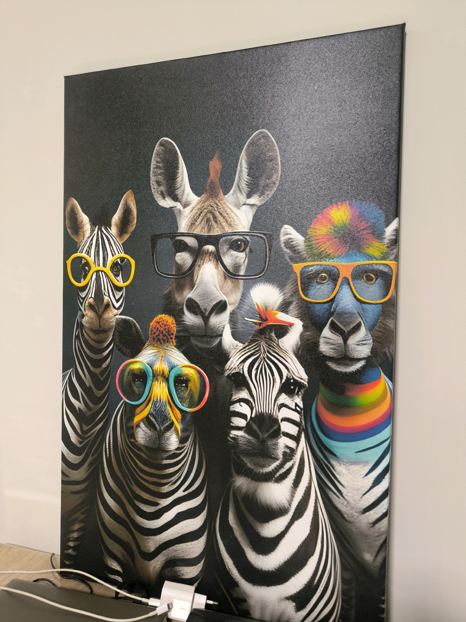 The image displays a framed canvas print on a wall, depicting a group of stylized animals wearing glasses. The animals include zebras, a giraffe, and what appears to be a llama or alpaca. Each animal has distinct accessories, such as brightly colored glasses, a rainbow-colored neckpiece, and a pom-pom on one zebra's head. The background of the print is dark and textured. In the foreground, at the bottom of the image, electrical cords and a white power adapter are visible on a dark surface, suggesting the canvas is hung near an electrical outlet. The wall beside the canvas is painted a light color. The scene is not set in a specific location in Magnitogorsk, Russia, as there are no identifying landmarks or contextual clues within the image to suggest this.
