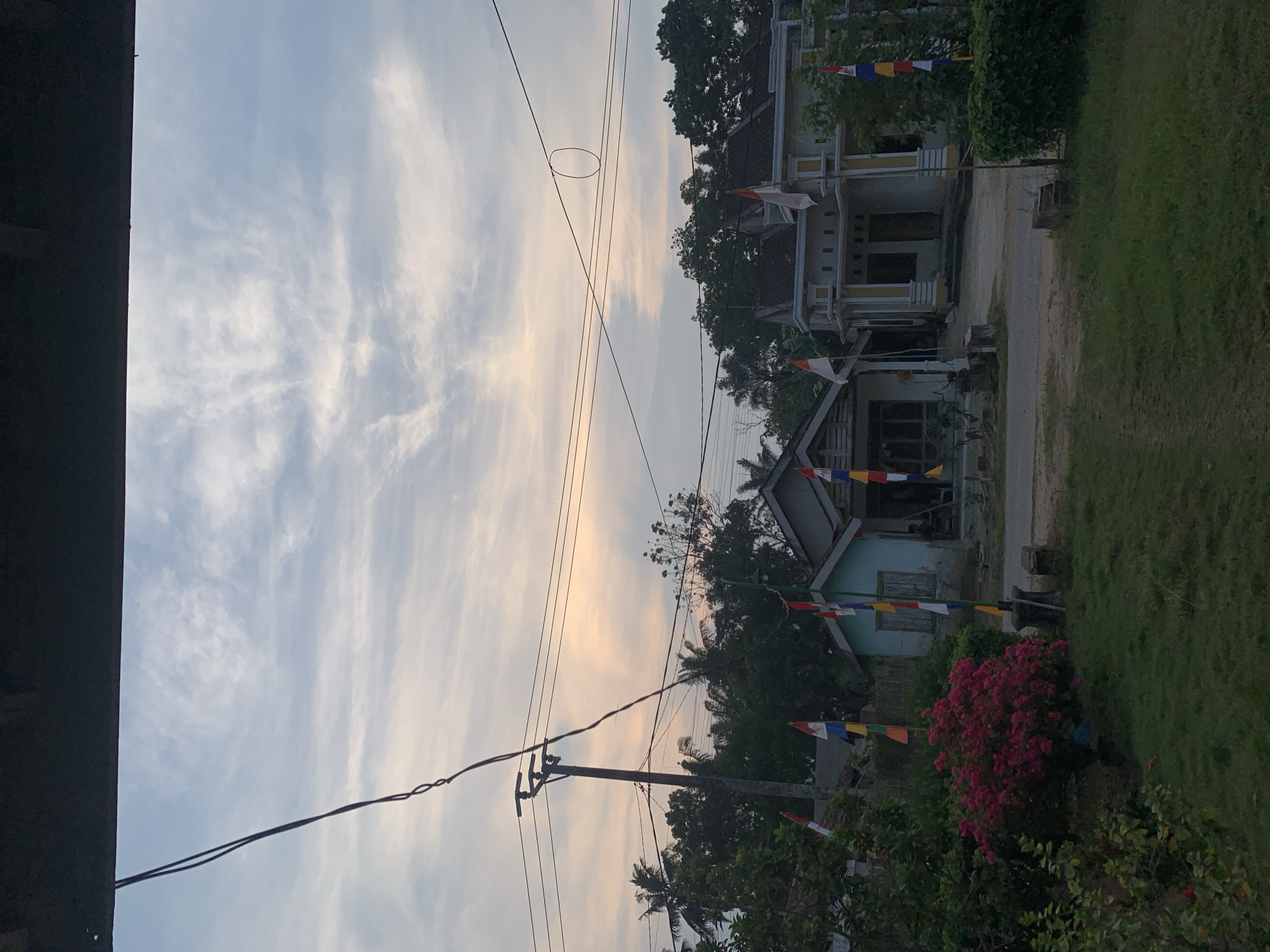 The image depicts a residential street scene in Rejo Asri, Indonesia, during what appears to be either sunrise or sunset, given the soft, warm light illuminating the sky and the clouds. In the foreground, a patch of green grass leads to a dirt road. On the right side of the road, a two-story house with a yellow and white facade is visible, adorned with flags. To the left of this house is a smaller, light blue house. Both houses are surrounded by lush green trees and vegetation. Colorful flags, likely representing the Indonesian national colors (red and white) and possibly other festive decorations, are planted on poles in front of the houses. A prominent utility pole with electrical wires stretches across the upper portion of the image, with a wire forming a rough circular loop. The sky is filled with wispy clouds, with streaks of orange and yellow light suggesting the low angle of the sun. There is one individual visible sitting on a chair in front of the blue house.