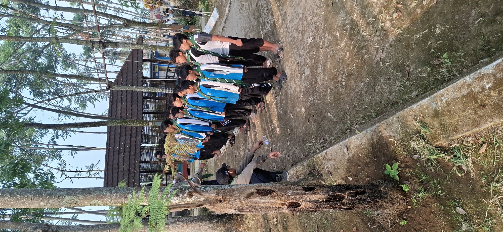 A group of approximately 15 boys, appearing to be of scouting age, are lined up in formation outdoors. They are dressed in blue and white scout uniforms, complete with green neckerchiefs. One individual, a scout leader, is kneeling on the left side of the frame, facing the line of boys and holding a small clear plastic cup. The environment appears to be a park or recreational area with trees, a dirt path, and a low concrete border. In the background, a wooden building with a tiled roof is visible, along with some ropes and structures suggesting an adventure park. The lighting suggests it is daytime, possibly morning or late afternoon. The location is identified as Puntukrejo, Indonesia.