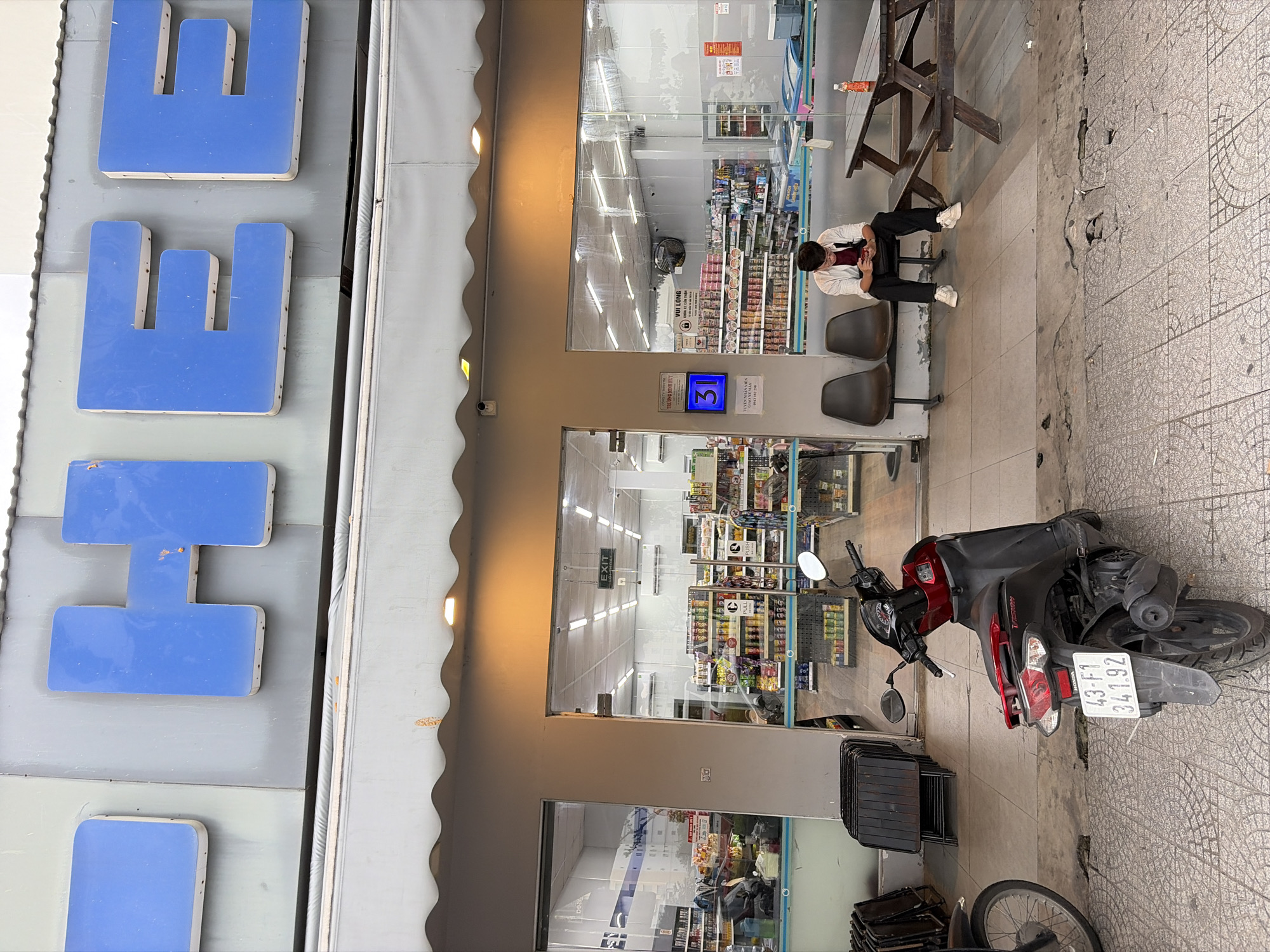 The image captures the exterior of a convenience store, likely a "Circle K" chain, on a bright day in Da Nang, Vietnam. The prominent blue sign above the entrance spells out "CHEE" in large, three-dimensional letters. Two chairs are situated outside the store, with a young man sitting on one, engrossed in his smartphone. A wooden picnic table with a bottle on it is to his right. In front of the store, a red and black scooter is parked, displaying a license plate that reads "43-F1 341.92". The pavement is tiled with a geometric pattern. The store entrance is wide and glass-fronted, revealing rows of shelves stocked with various products. A sign with the number "31" is visible to the left of the entrance, possibly indicating a queue number or a promotion. The overall scene suggests a typical urban street view in Da Nang, with a focus on a commercial establishment and its immediate surroundings.