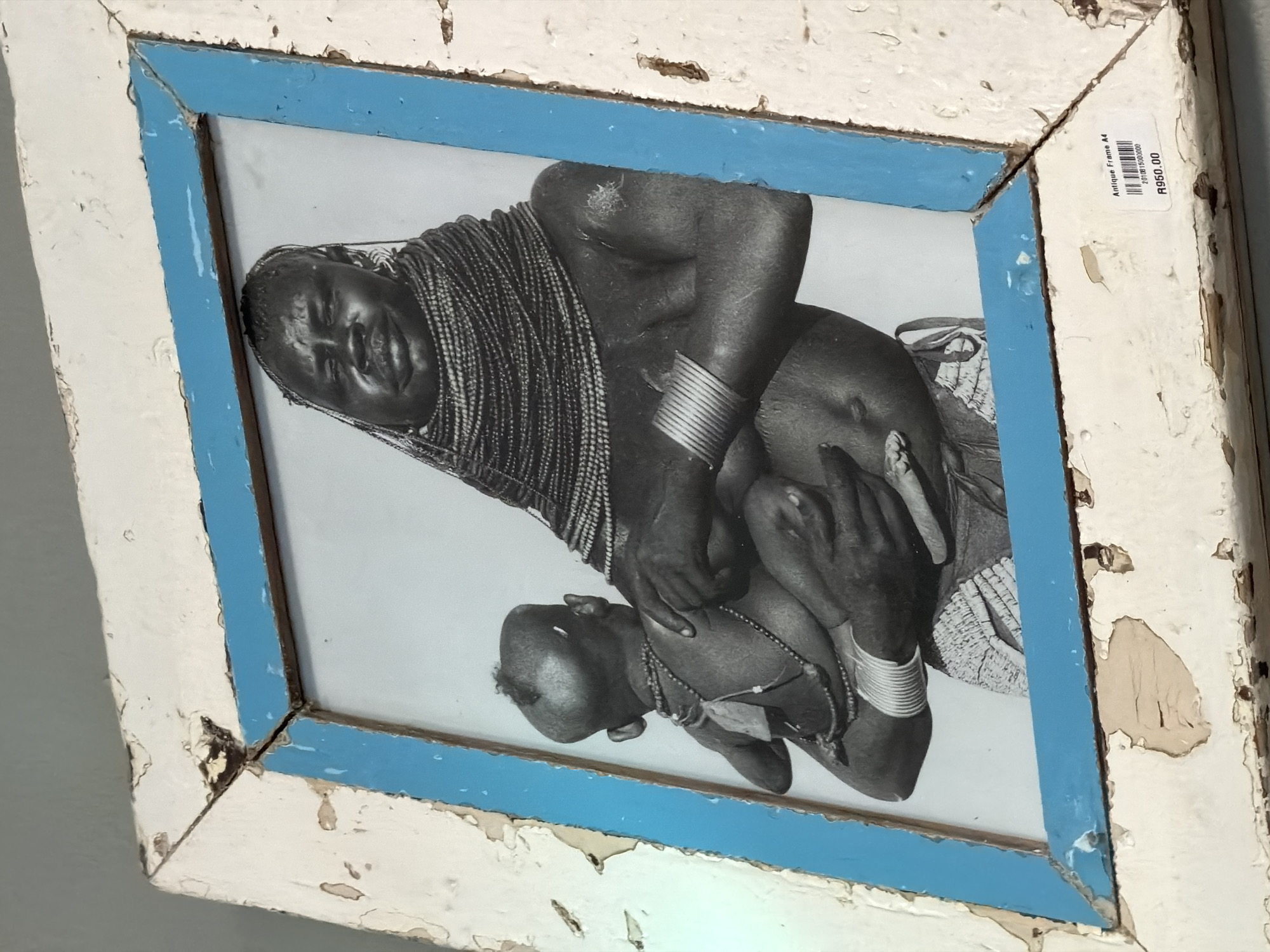 A black and white photograph, depicting an African mother and child, is showcased within a distressed frame, tilted against a light-colored wall. The photograph captures an intimate moment, with the adult figure, possibly the mother, holding the child. The adult has intricately braided hair and wears multiple ringed armbands. Her expression appears gentle and serene. The child, held close, wears a simple beaded necklace.
The frame itself is notable for its aged appearance. The outer section is painted a chipped and peeling white or cream, revealing underlying material. An inner border around the photograph is painted light blue, also showing signs of wear and flaking.
In the top right corner of the frame, a white sticker is attached. Visible text on the sticker reads "Antique Frame A4," followed by a barcode "7101815000000," and a price of "R950.00," indicating South African Rand. The distressed aesthetic of the frame and the price tag suggest the item may be for sale in a shop or market, consistent with the Cape Town location context.