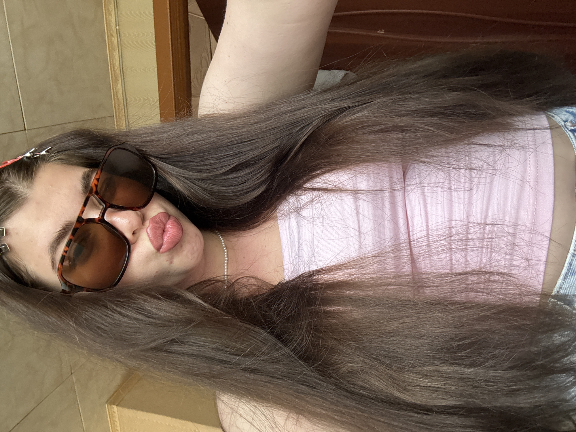 A woman with long brown hair is wearing sunglasses and a light pink top. The background includes a partially visible wooden structure. It is not possible to determine the location as Мещёры, Russia, from the image alone.
