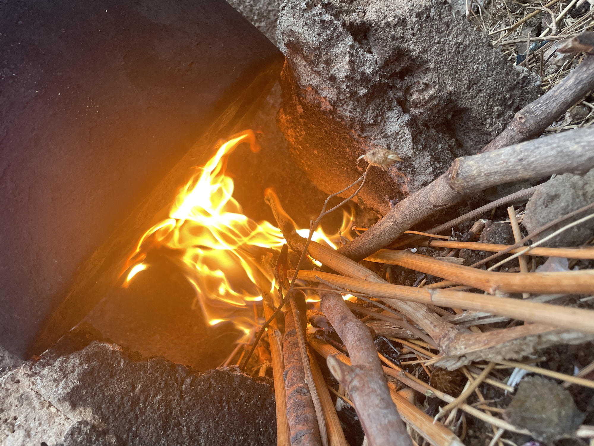 A fire burns inside a makeshift hearth constructed from a dark metal container and rough, gray stones. The fire consumes thin, dry branches that have been placed within the hearth. Outside of the immediate area of the fire, the ground is covered in scattered dry vegetation and dirt. The location is Jos, Nigeria.