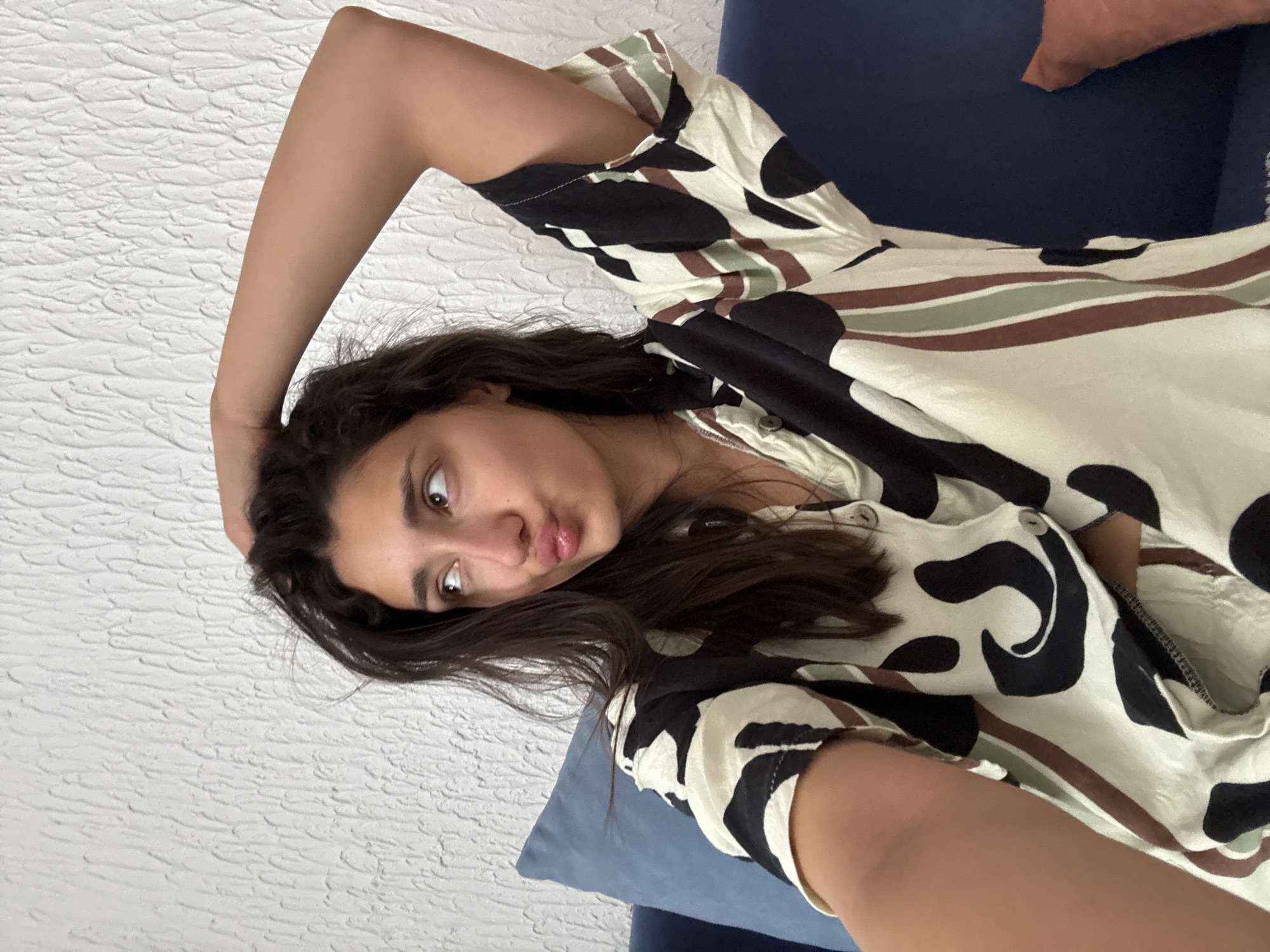 A woman is posing for a selfie, her head tilted to her left, arm raised and hand in her hair. She's wearing a patterned top with a buttoned front. She is lying on a blue cushion, perhaps a sofa. The background includes a textured white wall. There is no indication of Sutomore, Montenegro, or any city location.