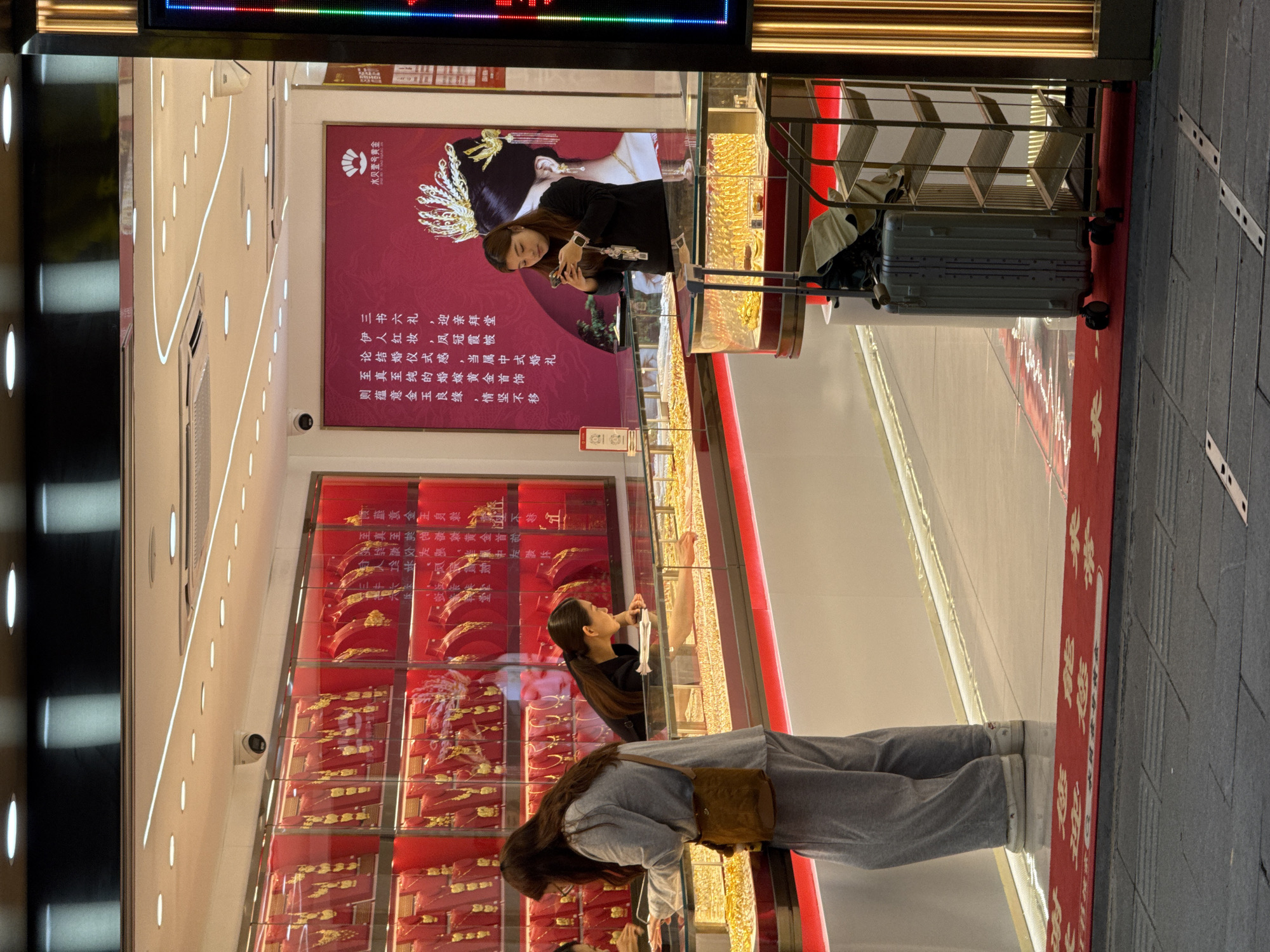 Here is a description of the image:
The photograph depicts the interior of a jewelry store, with two women looking at displayed gold jewelry. One woman, in a gray outfit, is peering closely into the display, while the other, in black, is taking a picture. A suitcase with a beige coat rests behind the woman taking the picture, outside the store. A large poster with Chinese text hangs inside the store promoting a “Water Bay Number One Gold." The exterior flooring consists of grey stone pavers. The jewelry store is located in Liwan District, China.