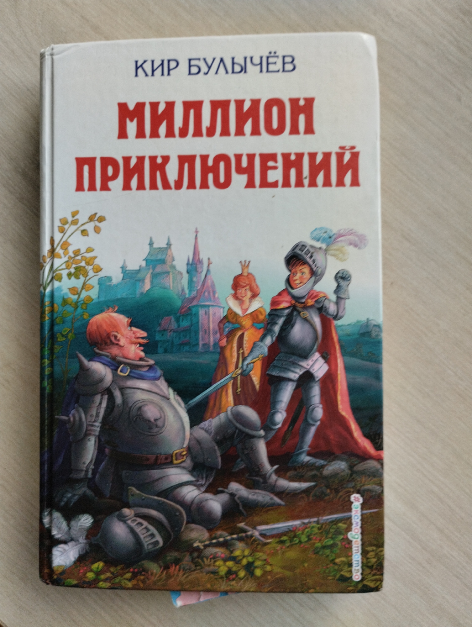 A book cover is pictured lying on a wooden surface. The book is titled "Миллион приключений" (A Million Adventures) by Кир Булычёв (Kir Bulychev). The cover illustration depicts a medieval scene with several characters. In the foreground, a knight in armor is kneeling or lying on the ground, while another knight stands triumphantly. A woman in a golden dress and crown is visible behind the standing knight, and a castle complex is rendered in the background. The overall setting appears to be a fantasy landscape. The image does not provide any information related to the city of Astravyets, Belarus.