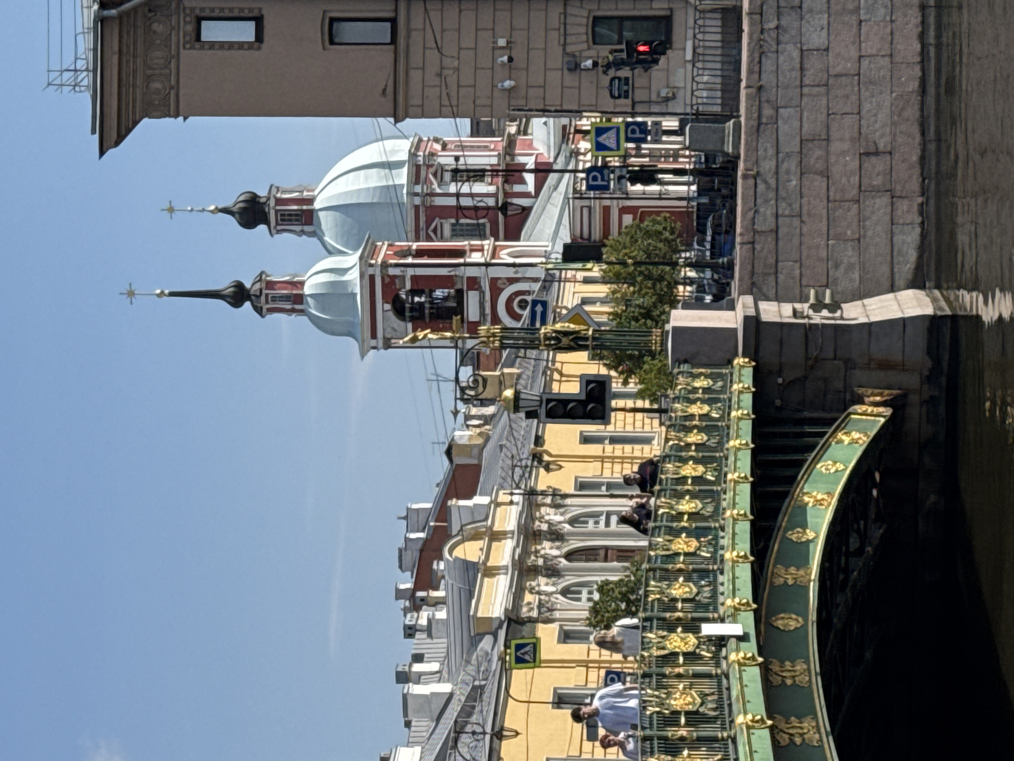 This is a photograph taken in Saint Petersburg, Russia, likely on a sunny day, featuring a view of a canal with a decorative bridge in the foreground. The bridge, painted green with gold ornamentation, spans the canal and has a stone embankment on the right side. Several people are visible walking across the bridge.
In the background, there are buildings with yellow facades and ornate architectural details. A prominent red and white domed church with spires is situated behind these buildings, against a clear blue sky. Traffic lights and street signs are visible on the street behind the bridge. The water of the canal reflects the surrounding structures and the sky. The overall scene depicts an urban landscape with historical architecture, characteristic of Saint Petersburg.