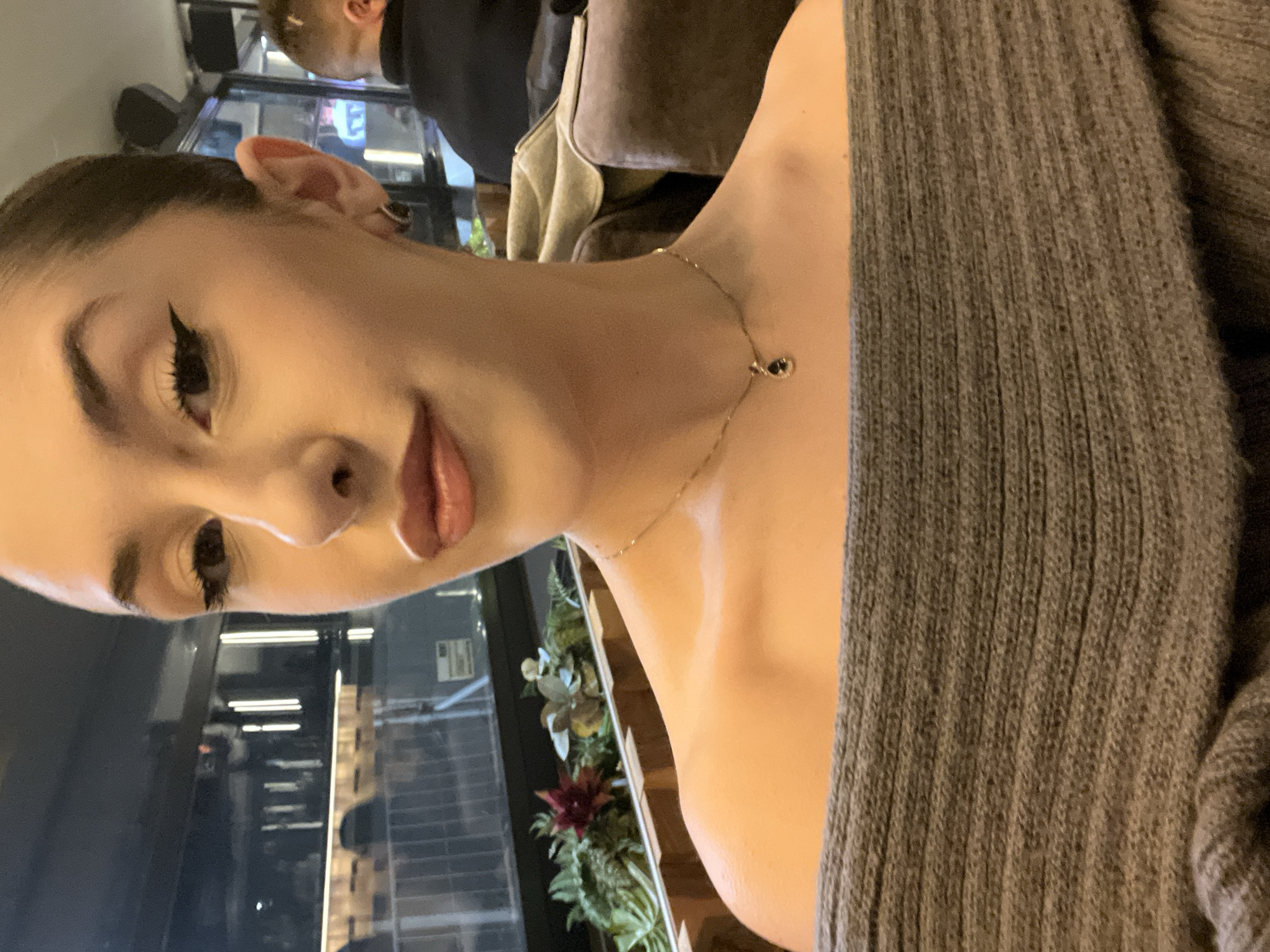 A young woman is posing for a selfie indoors, likely in a café or restaurant. She is wearing a grey, knitted sweater and a gold necklace with a dark pendant. Her makeup includes dark eyeliner and nude lipstick. In the background, a man is partially visible. The interior includes windows, plants, and furniture. The location appears to be in a modern, urban setting, potentially Sofia, Bulgaria.
