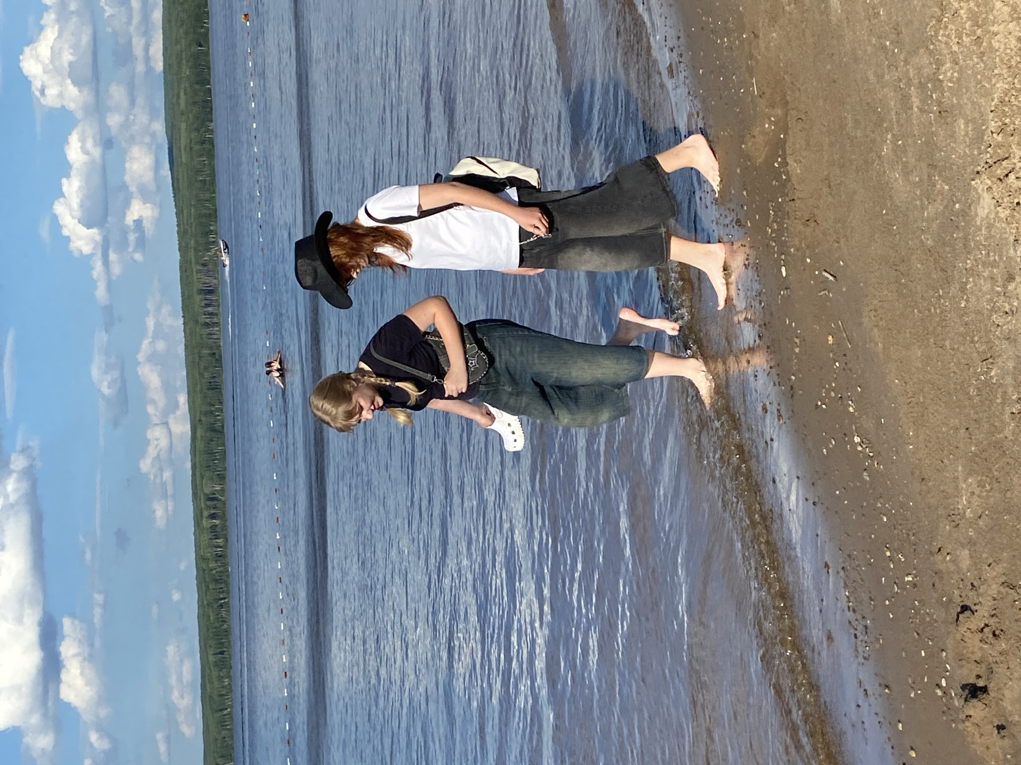 Two young women are walking barefoot along the sandy shore of the Kama River in Chaikovsky, Russia. The woman on the left has blonde hair in pigtails, wears a black t-shirt and denim capri pants, and carries a white shoe. The woman on the right has red hair, wears a white t-shirt, dark denim capri pants, and a black cowboy hat, and carries a backpack. They are walking towards the right side of the frame, with shallow water lapping at their feet. In the background, the river is calm, with a few boats and people visible on the water, and a treeline along the far bank. The sky is blue with scattered white clouds. The overall scene depicts a casual summer day at the beach.