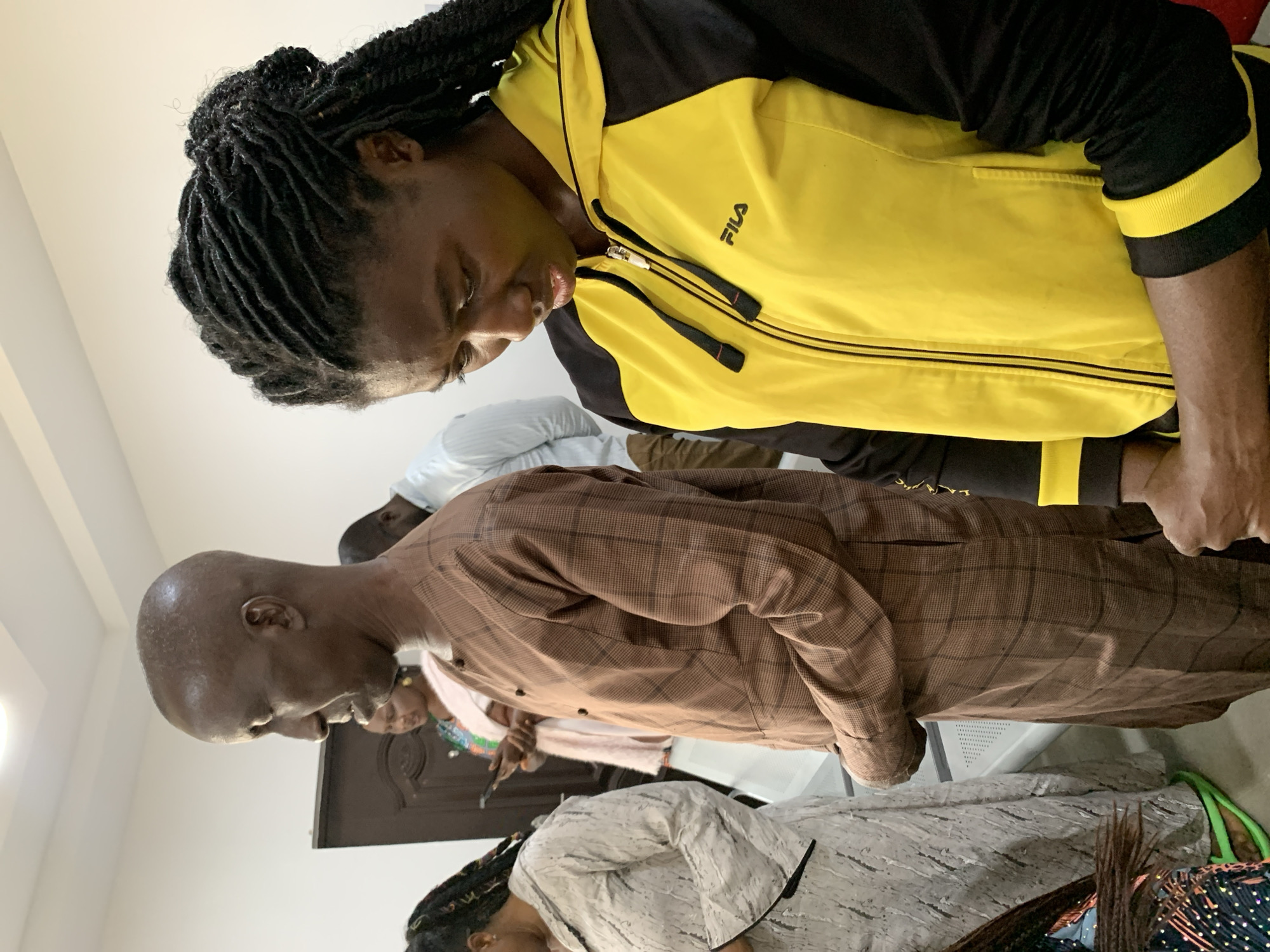 The image shows a group of people standing in an indoor setting, possibly a church or community hall, in Jos, Nigeria. In the foreground, a woman with dark, braided hair is wearing a yellow and black zip-up hoodie with "FILA" logo visible. Next to her, a man with a bald head is wearing a brown checkered kaftan. To the left of the man, another woman in a grey patterned dress is looking down at her phone. Further back, a man in a white shirt can be seen. The room appears to be simply furnished with white walls and a drop ceiling with fluorescent lighting. The overall atmosphere suggests a gathering or service.