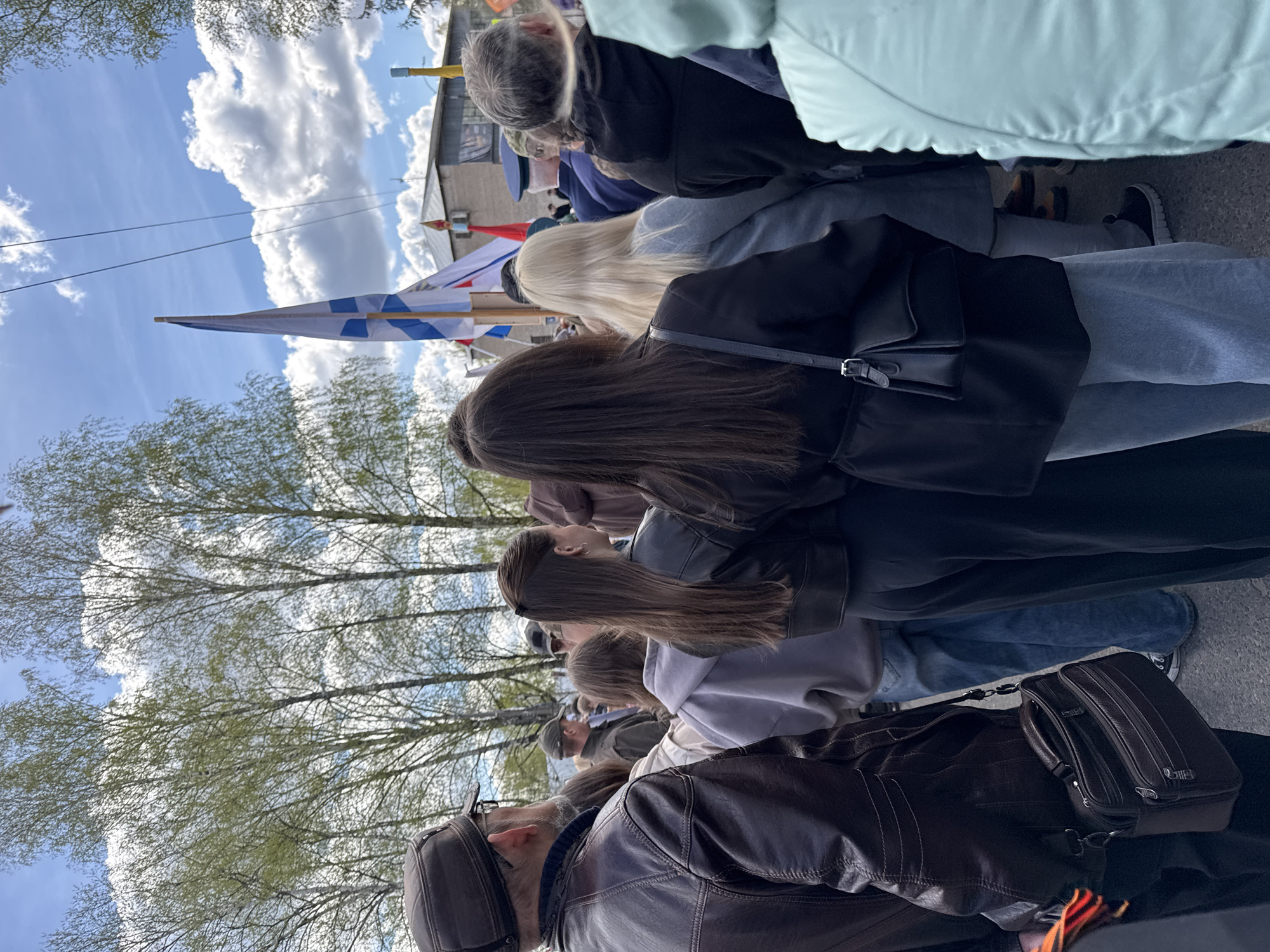 A crowd of people is standing outside in the city of Chuprino, Russia, during a daytime event. The sky above shows bright blue with scattered white clouds. There are several tall trees with light green leaves partially obscuring the view. In the background, there is a building with a yellow structure and a red object on top. The people in the crowd vary in age and attire. Several are wearing jackets, and some are carrying bags. A flag with a white background and blue stripes is visible, held up above the crowd.