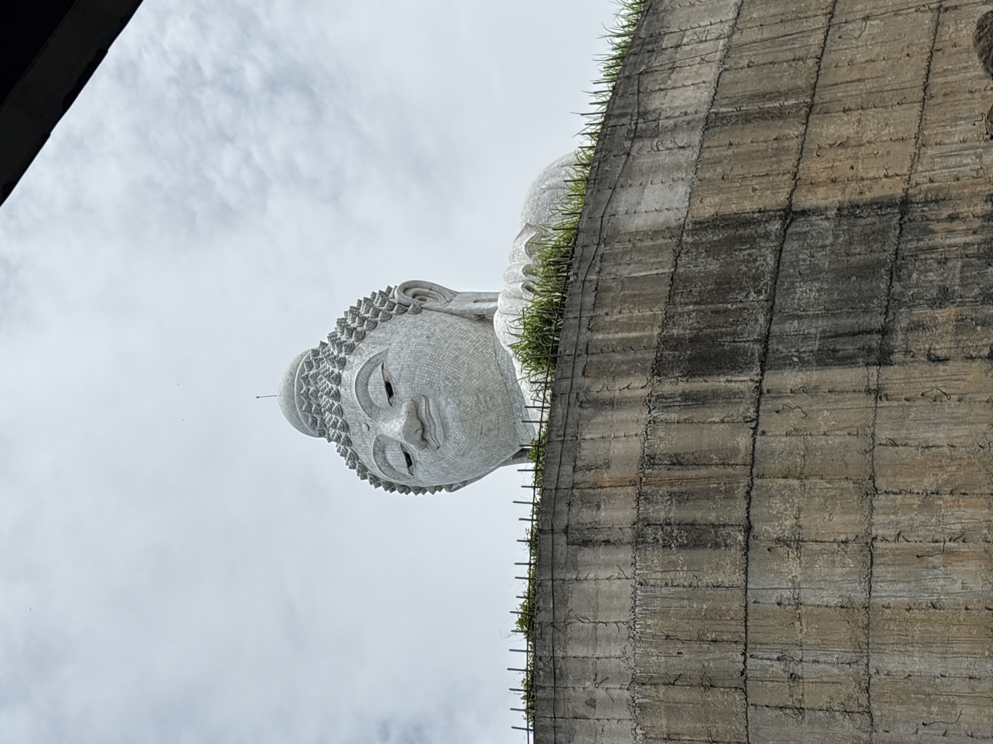 The image shows a low-angle, medium shot of the Big Buddha statue in Rawai, Thailand. The statue is made of white marble and is partially visible, with its head and shoulders dominating the frame. The statue is adorned with a patterned headdress and a serene expression. It is perched atop a concrete structure, which has a weathered, textured surface and is lined with green grass and a metal fence. The sky is overcast with light clouds. The overall impression is one of grandeur and peacefulness, with the weathered concrete base providing a stark contrast to the pristine white statue.