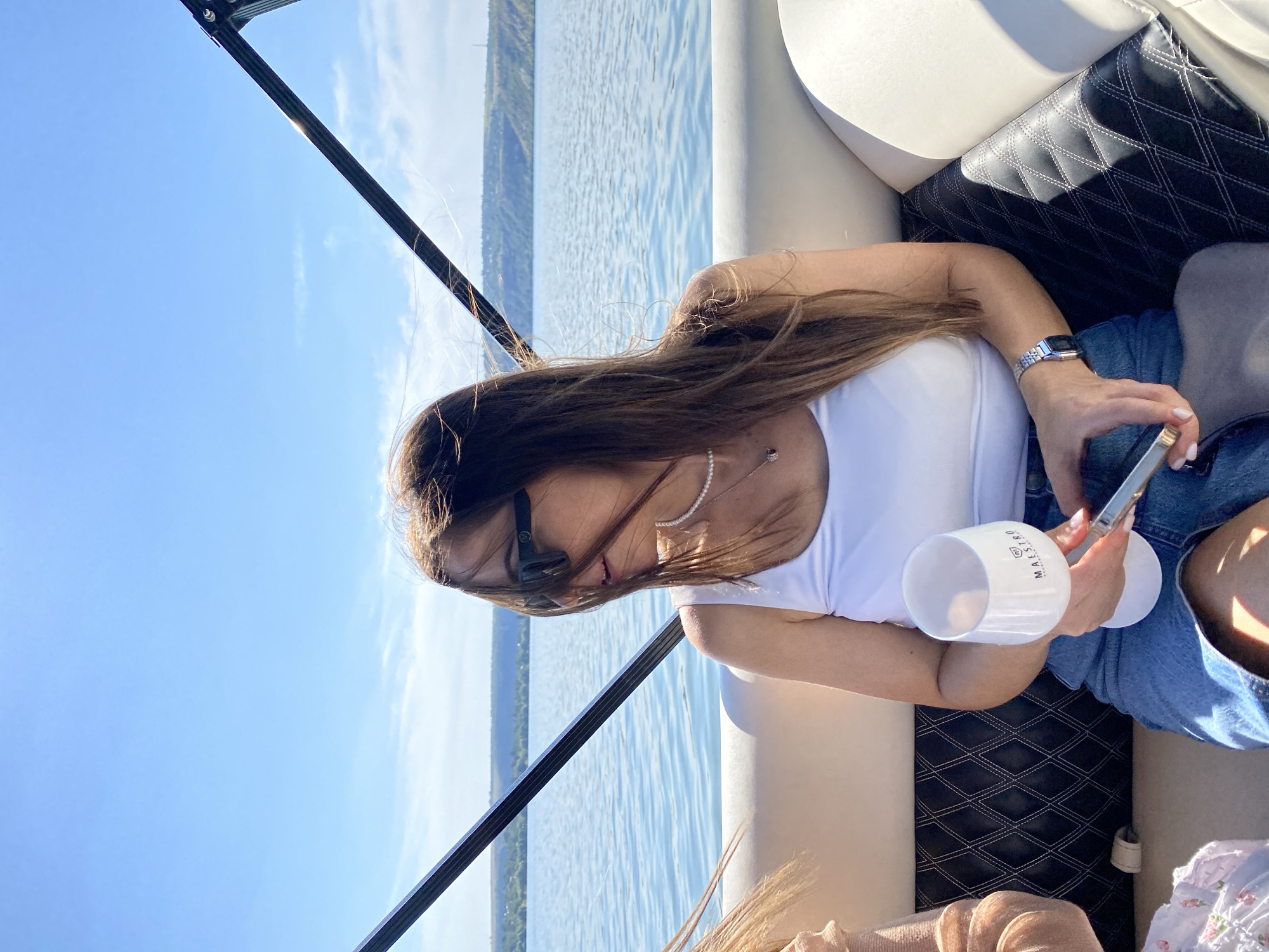 A woman with long brown hair, wearing sunglasses and a white tank top, sits on a boat in the city of Molovata, Moldova. She holds a wine glass in her right hand and a phone in her left. The boat is on a body of water with hills in the background. The sky is blue and clear.